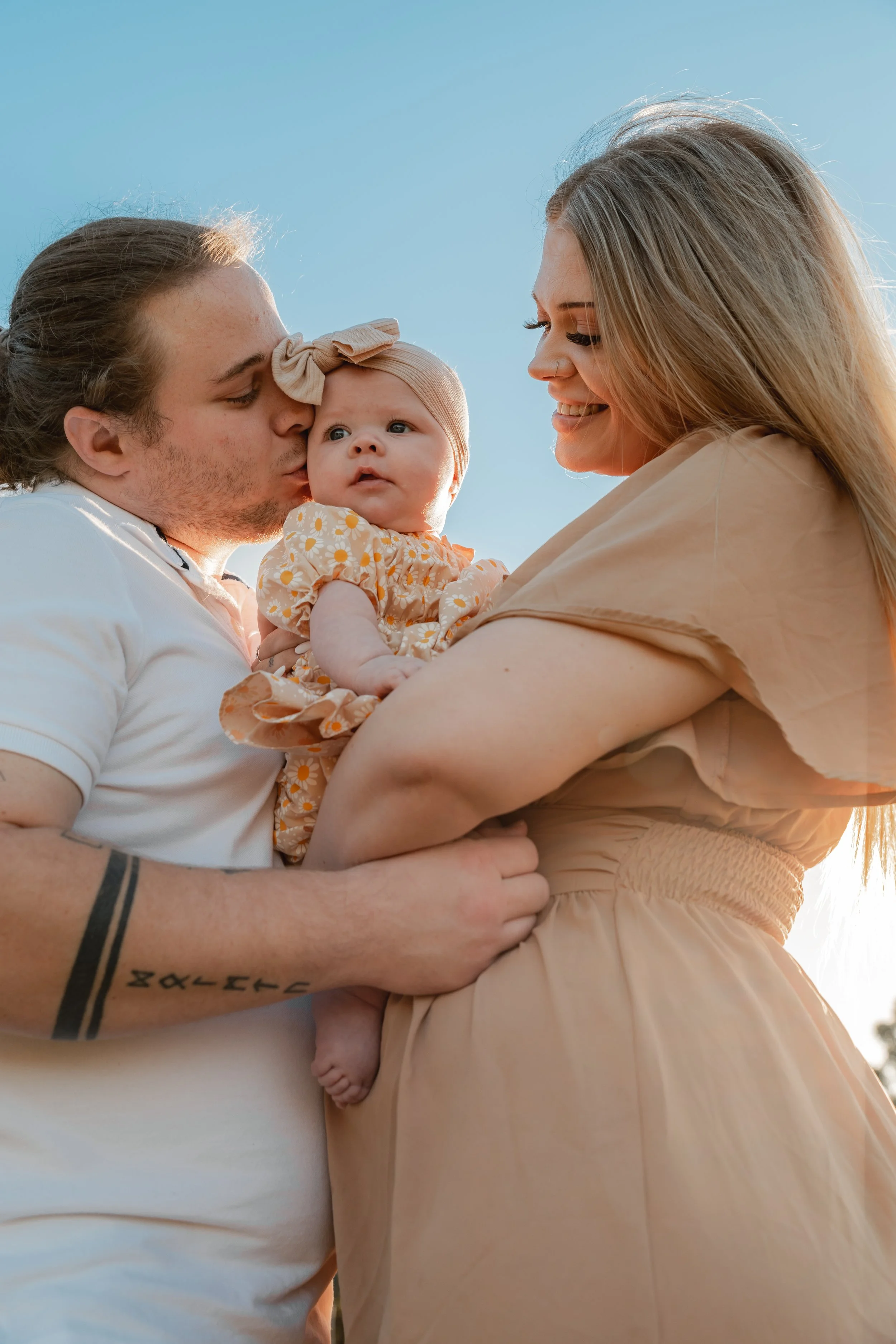 A family of three, a man, woman, and a baby girl, outdoors during sunset. The man and woman are holding the baby, and the man is kissing her cheek. The woman is smiling at the baby. The sky is clear with a light blue color.