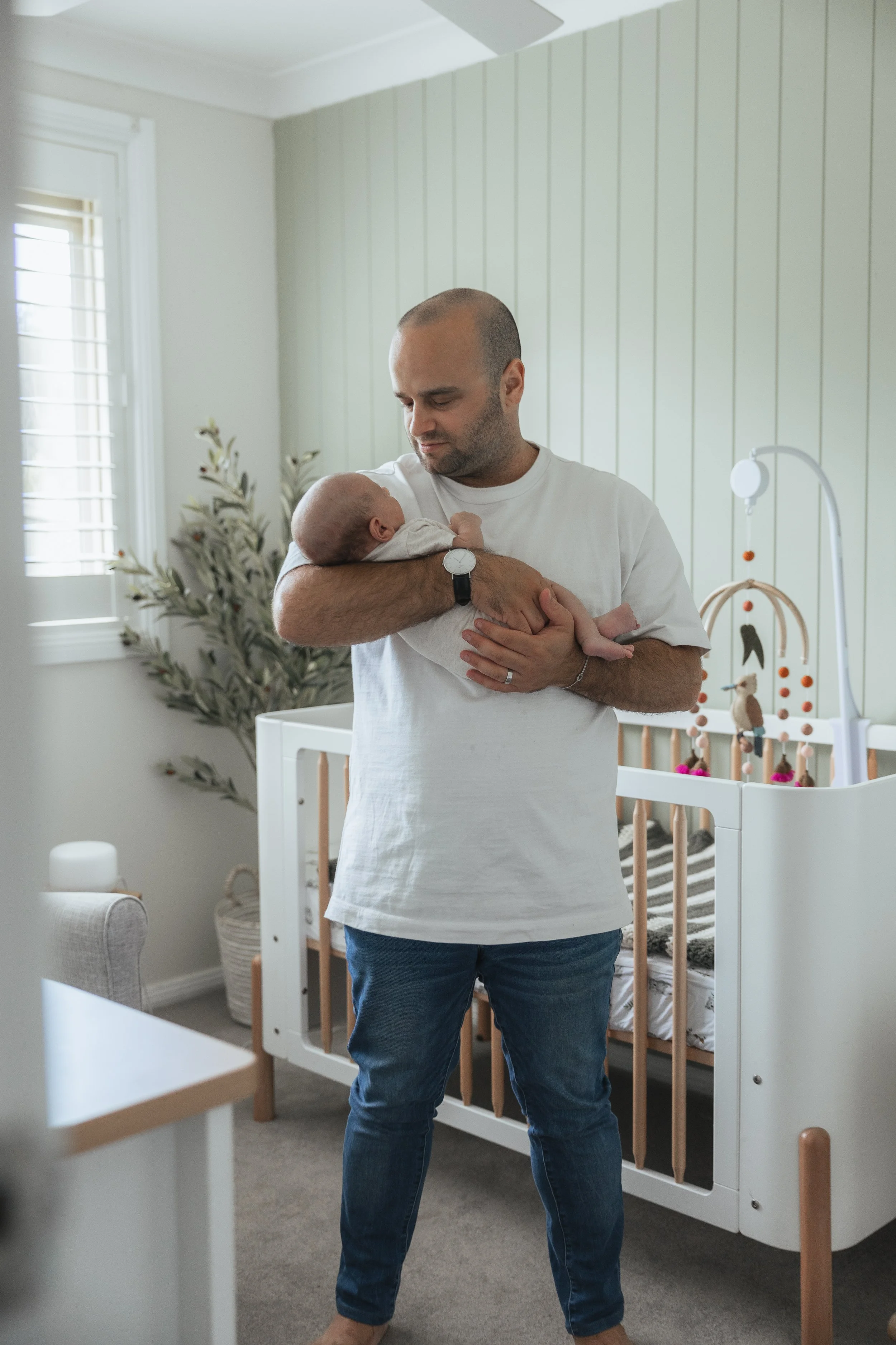 A man holding a baby in a nursery, standing next to a crib with toys, a potted plant, and a window with blinds.