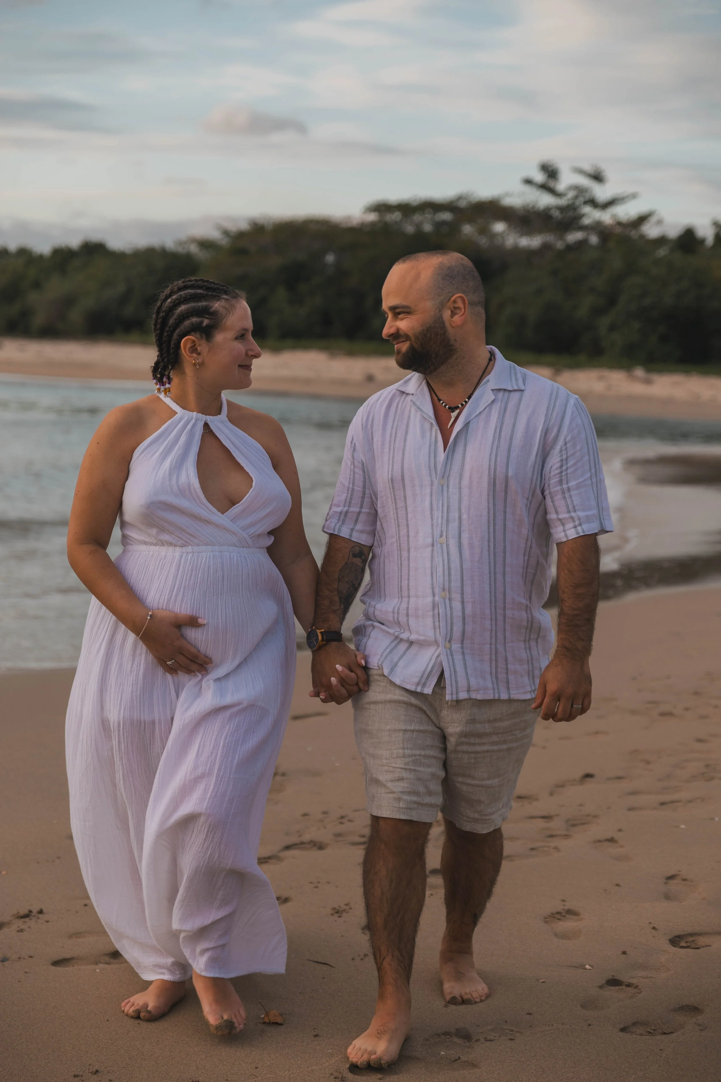 A pregnant woman and a man holding hands and walking on the beach at sunset, looking at each other.