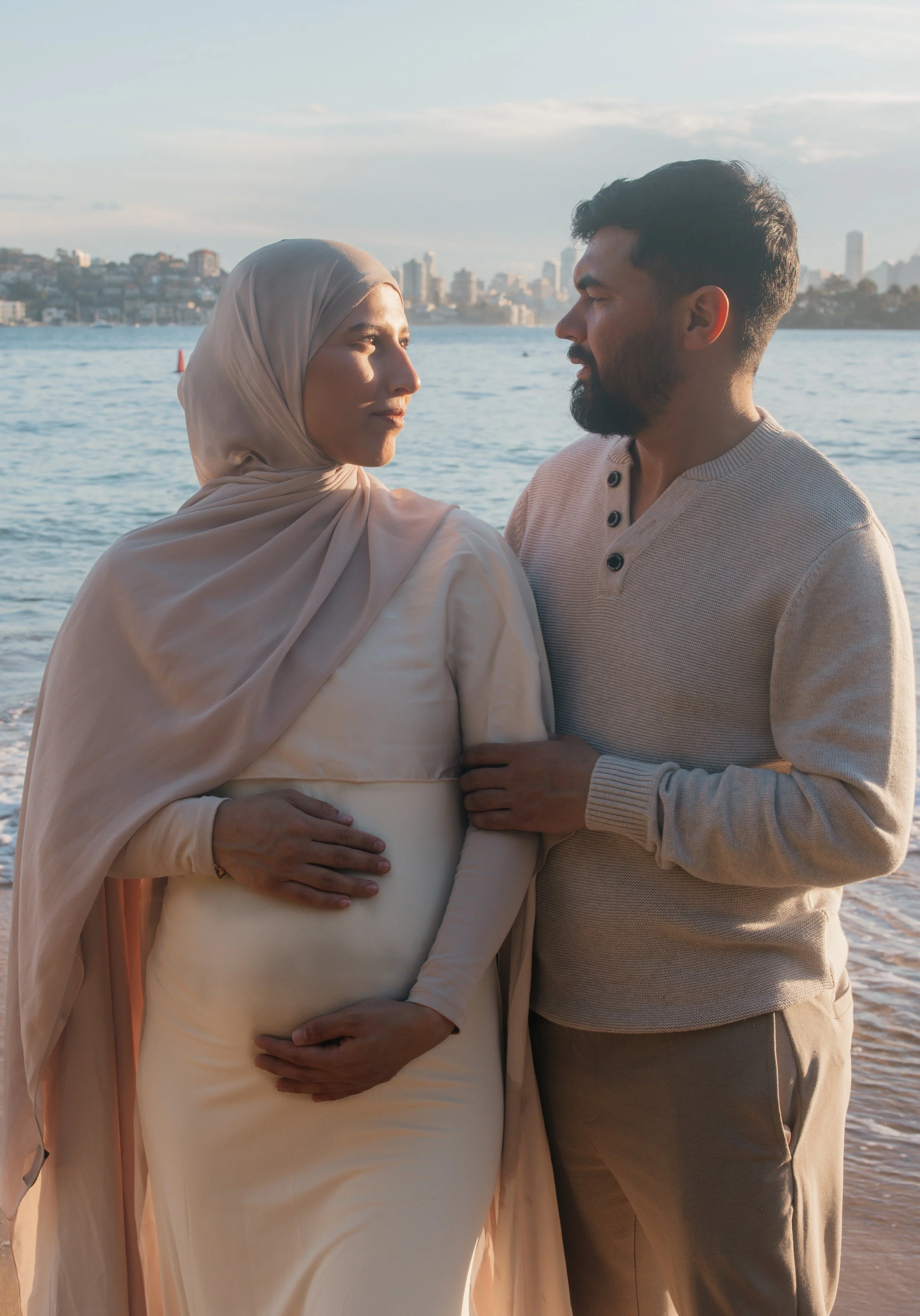 A pregnant woman wearing a beige hijab and dress standing on the beach holding her belly, while a man in a beige sweater stands close to her with his hand on her arm, looking into each other's eyes with a city skyline in the background during sunset.