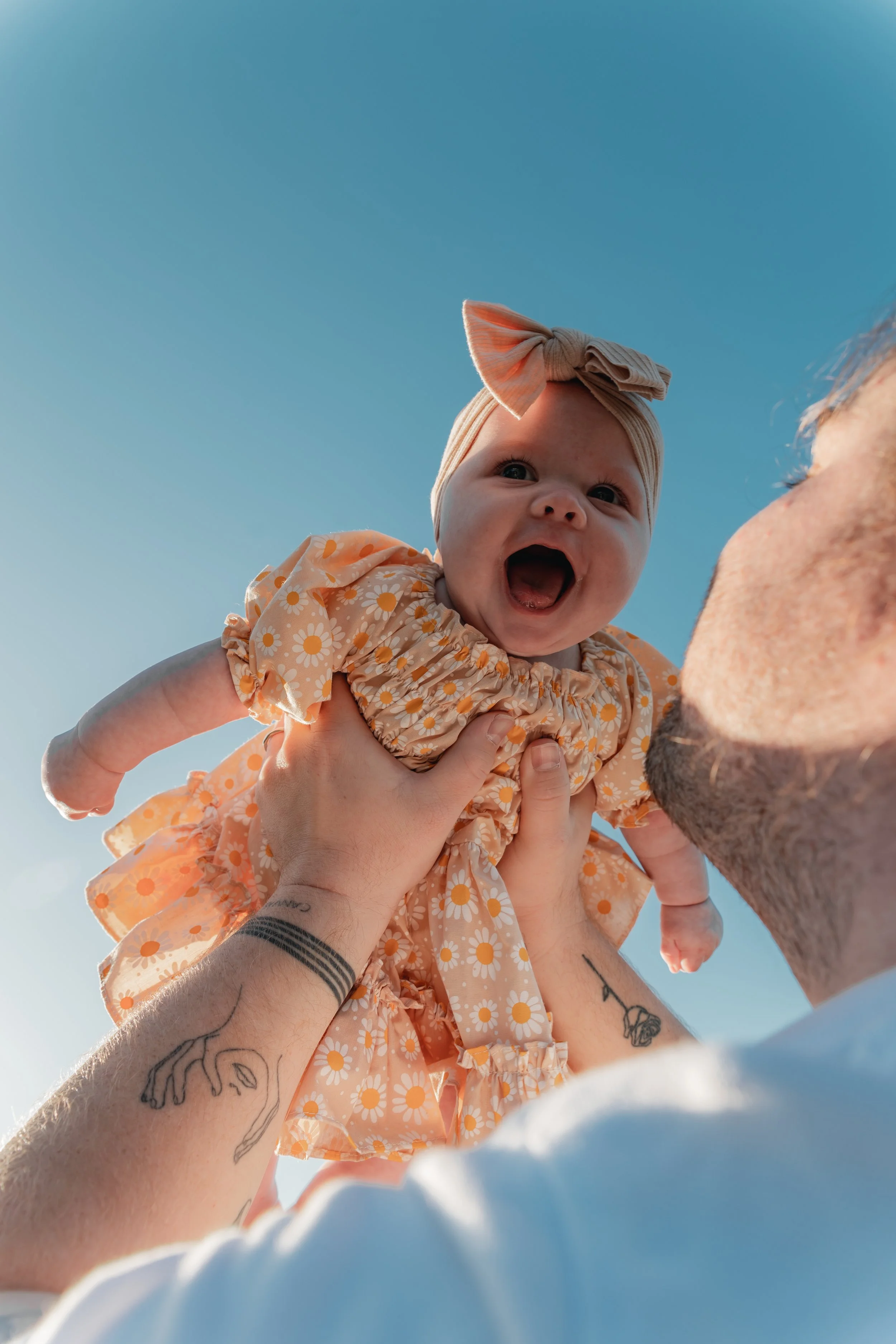 A man lifting a smiling baby girl into the air during daytime outdoor under clear blue sky.
