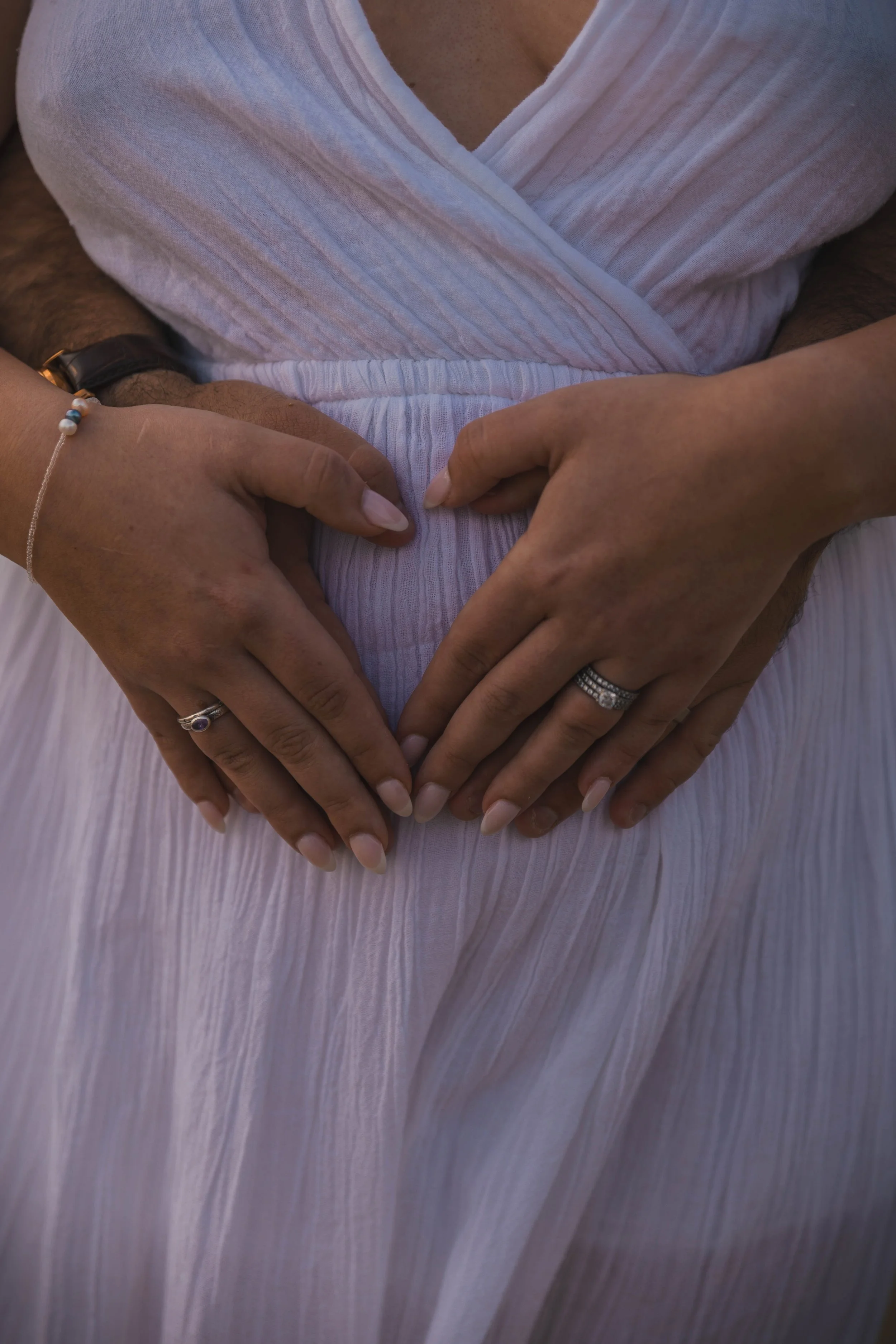 Two people hold their hands over a pregnant belly, forming a heart shape. One person wears a ring and a bracelet, while the other wears a ring, both with light-colored nails. The person is wearing a white, pleated dress.