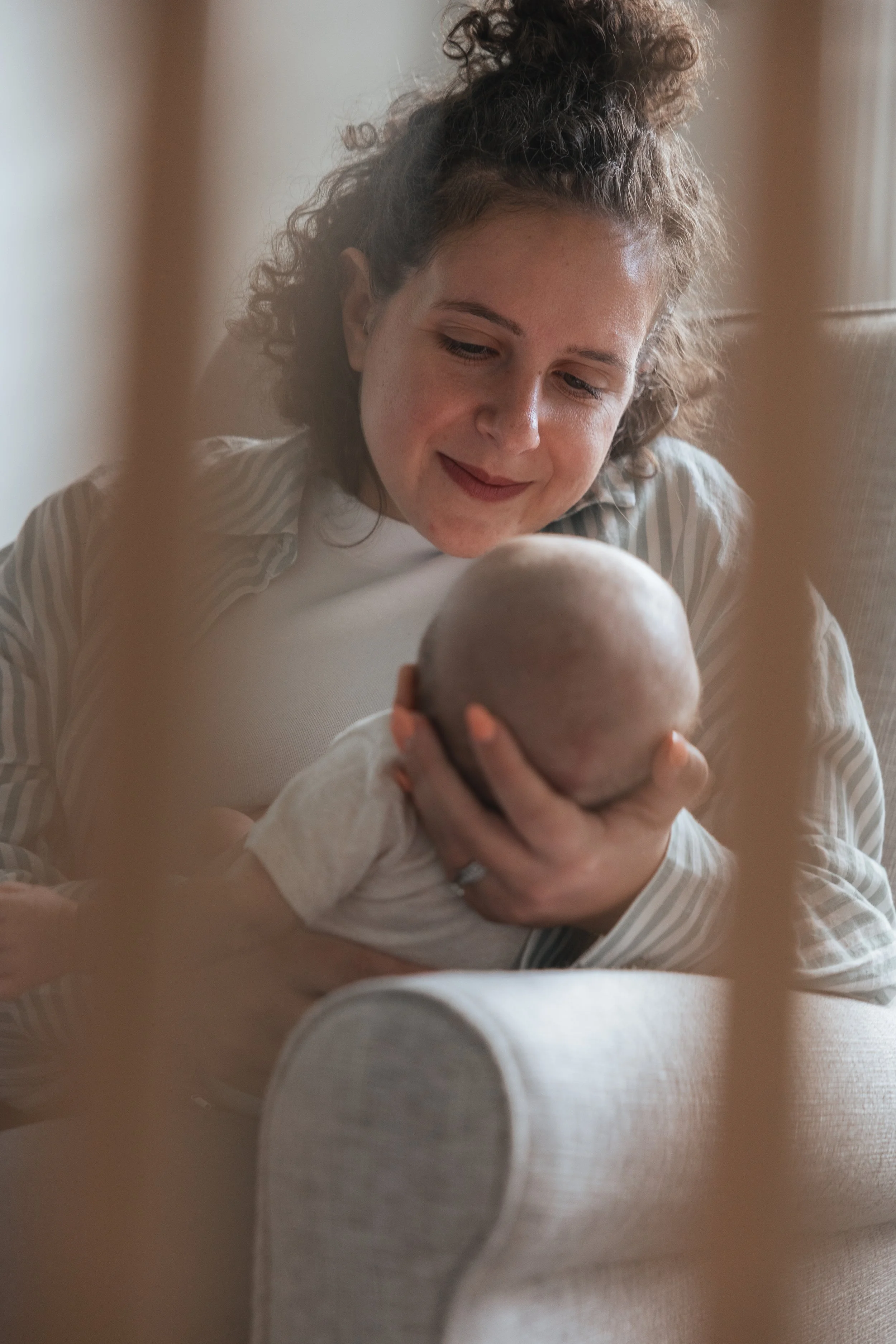 A woman with curly hair and a striped shirt looks fondly at a baby she is holding, seen through a blurred frame.