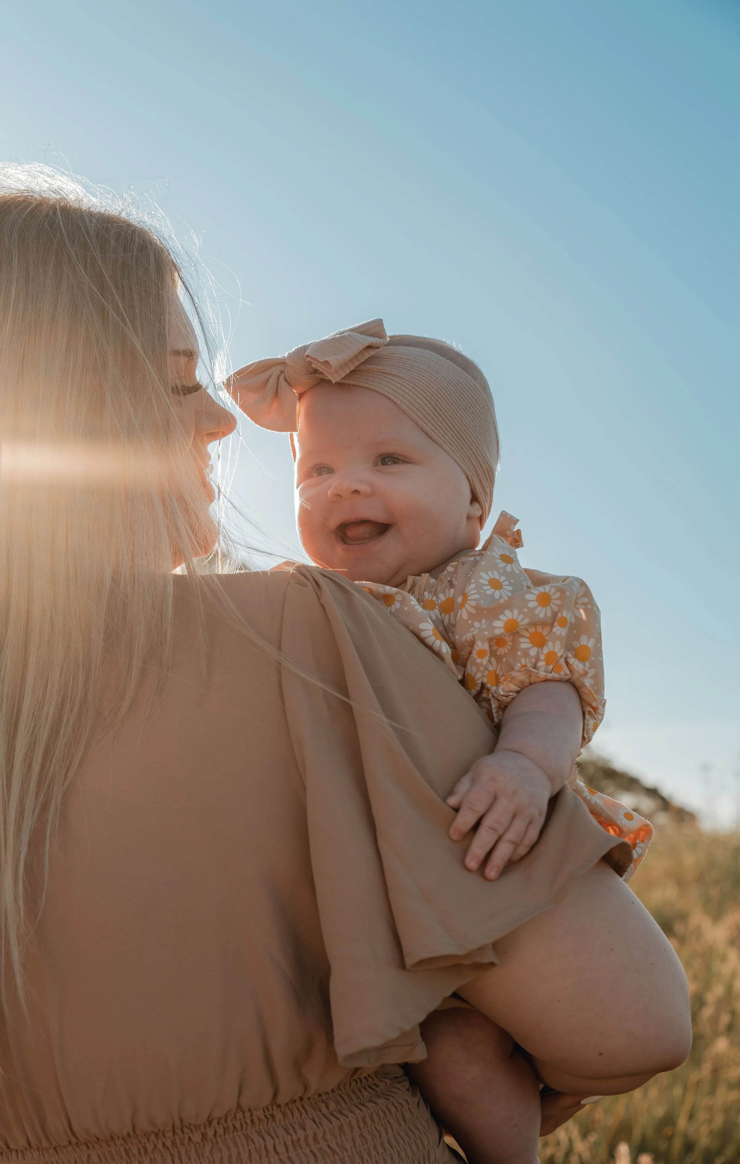 A woman holding a smiling baby girl outdoors in sunlight with clear blue sky.