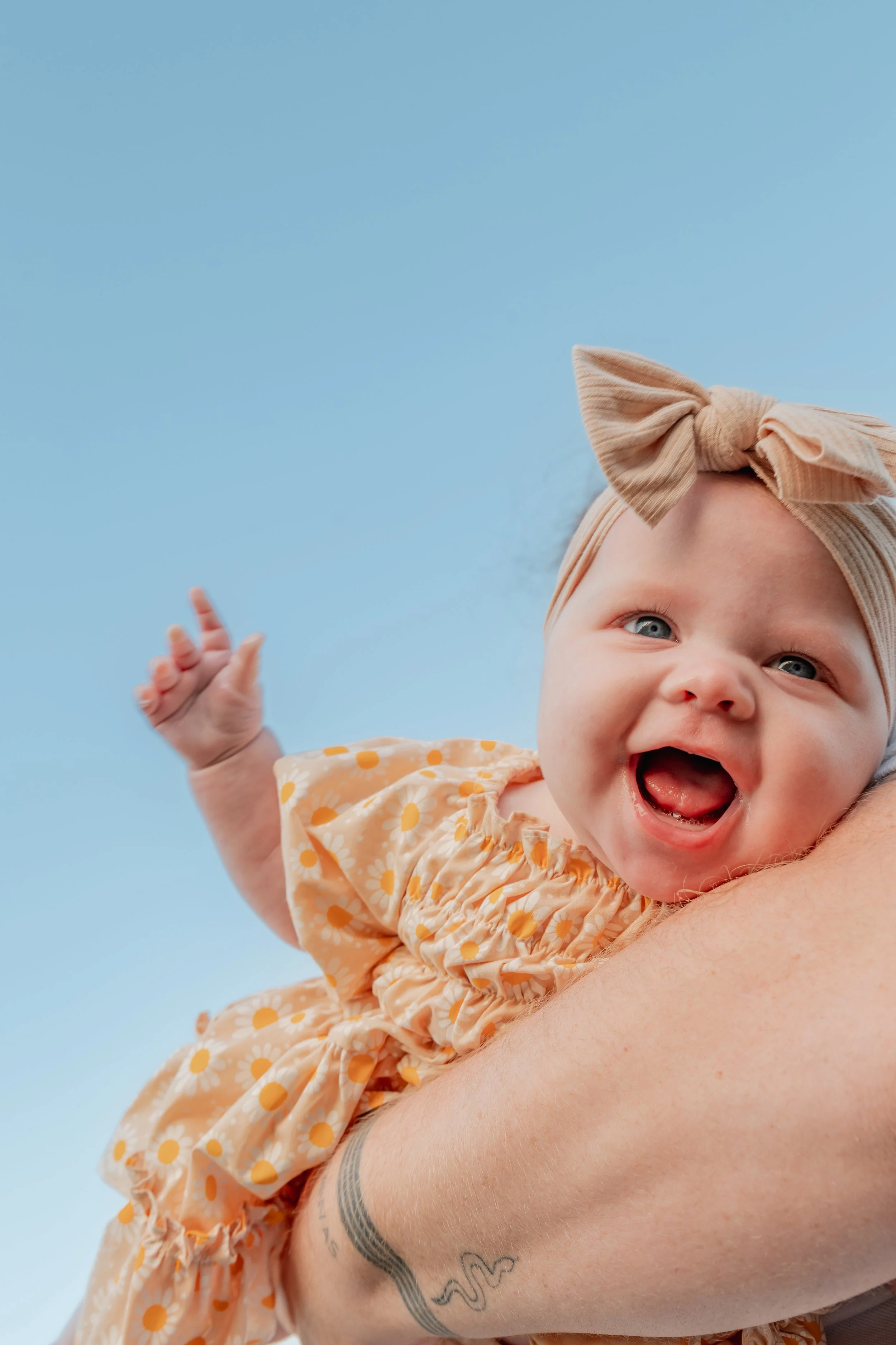 A smiling baby girl with blue eyes wearing a beige headband with a bow and a yellow dress with white daisies, being held up outdoors against a clear blue sky.