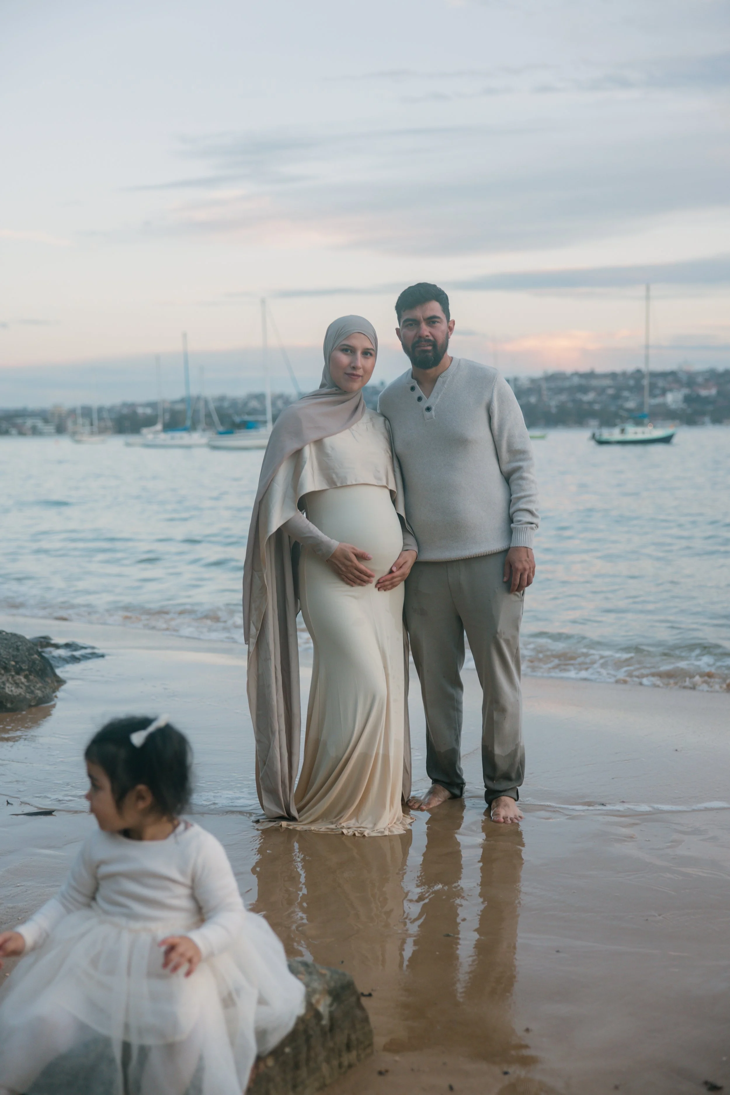A pregnant woman and a man standing on the beach near the water, with a young girl sitting on a rock in the foreground. The woman wears a headscarf and a long dress, and the man wears a sweater and pants. Sailboats are visible on the water in the bac