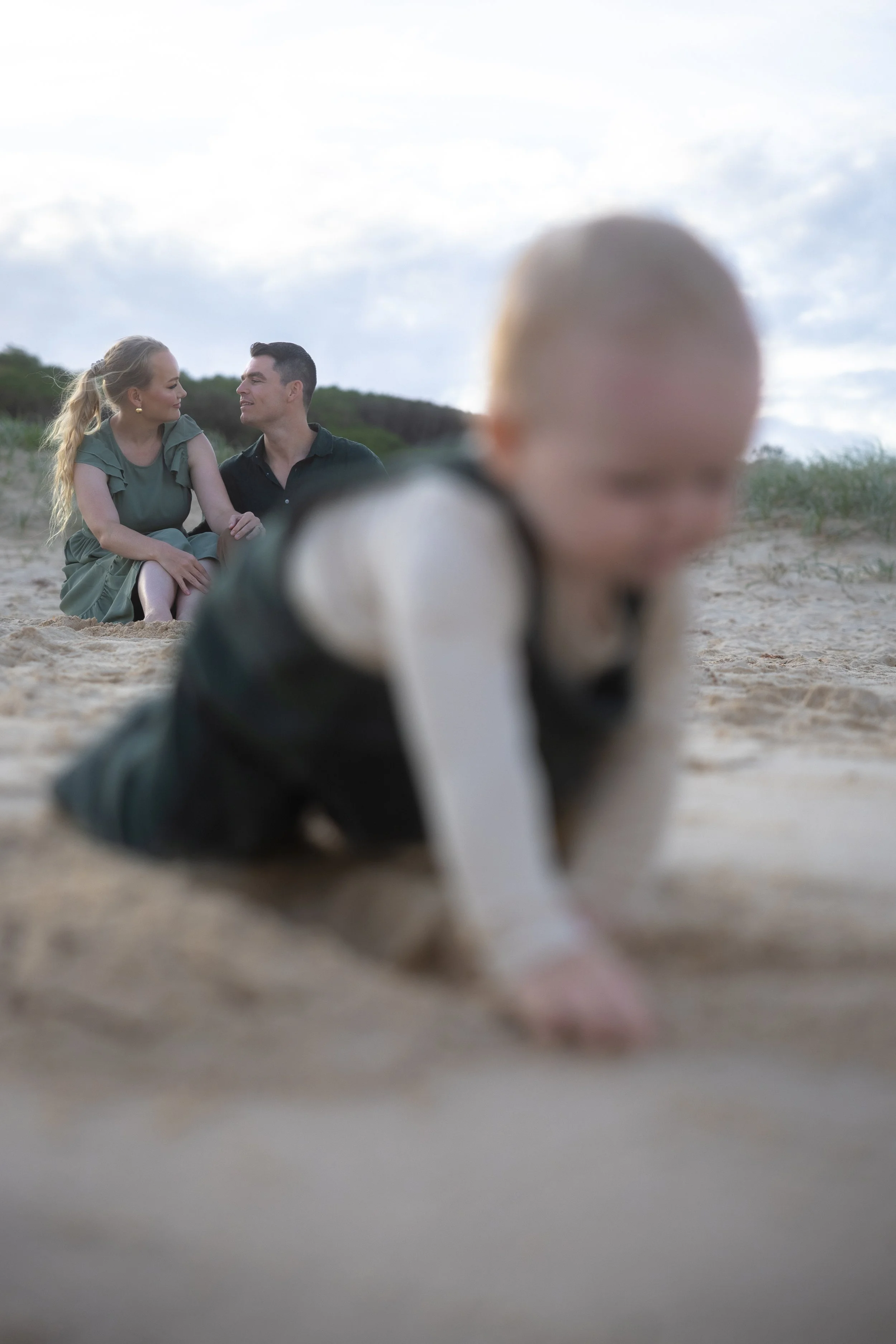 A young child crawling on the sandy beach in the foreground, with a couple sitting on the sand in the background, looking at each other, with a cloudy sky overhead.