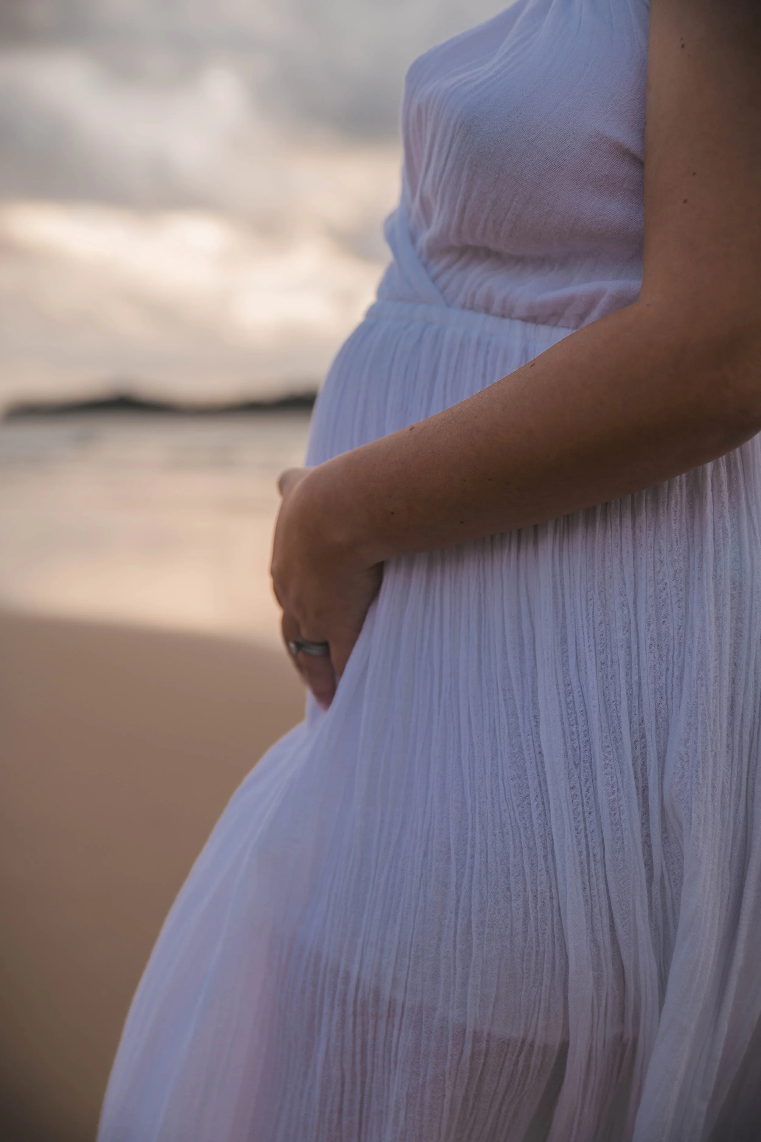 Close-up of a pregnant woman in a white dress on the beach with ocean waves in the background.