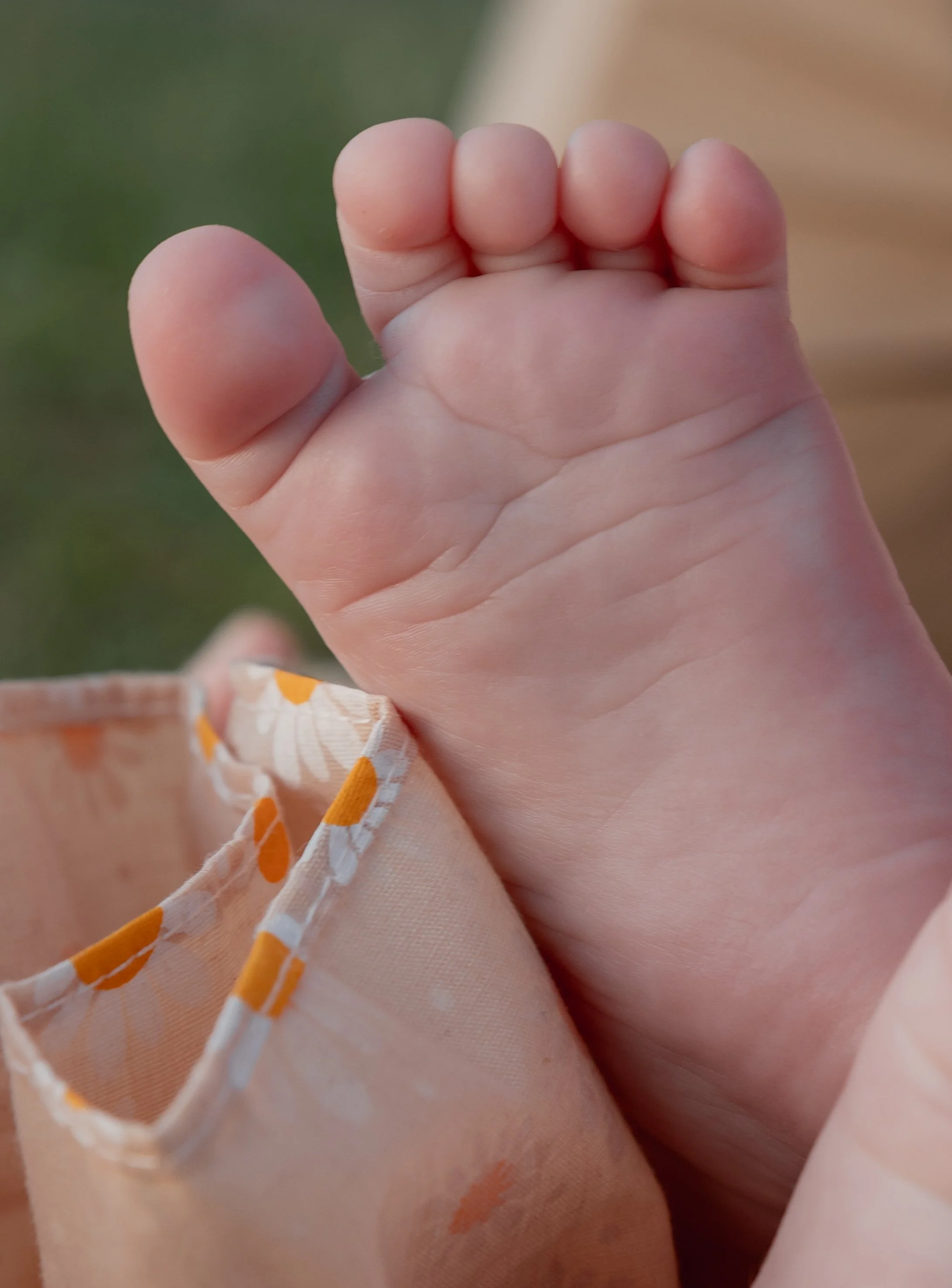 Close-up of a tiny baby's hand gripping fabric with orange and white patterns, outdoors with blurred green background.