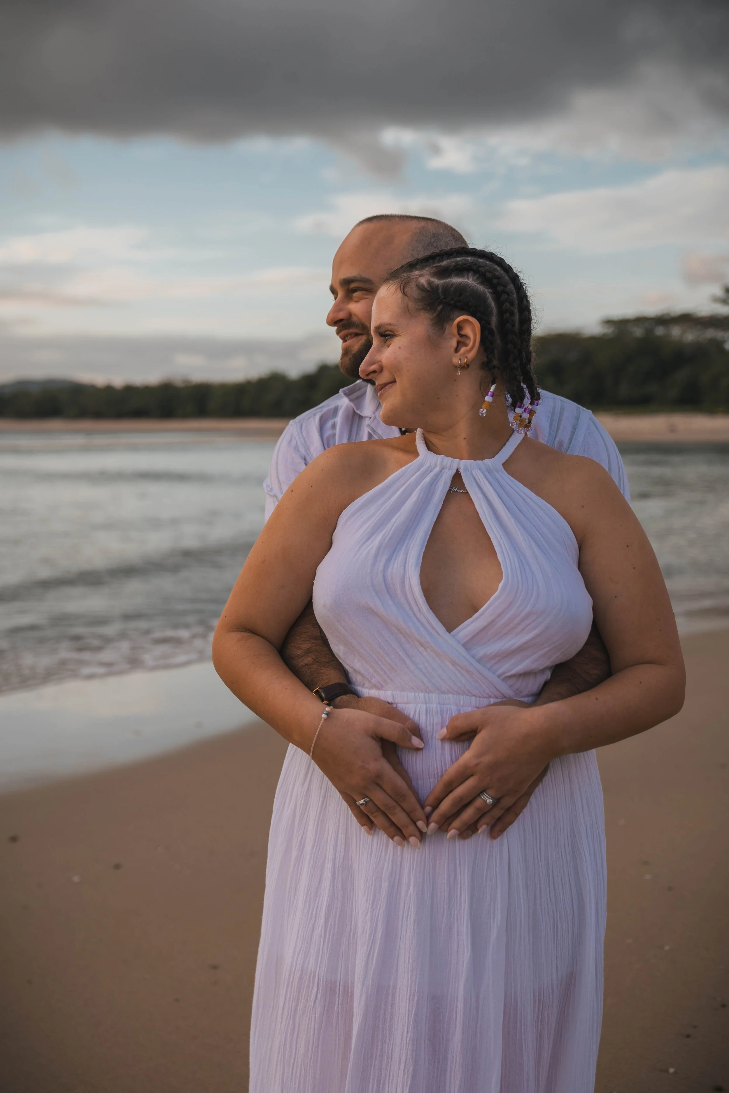 A couple stands on the beach, embracing and smiling, with the man's arms around the woman's waist and her hands resting on her stomach, under a cloudy sky near the shoreline.