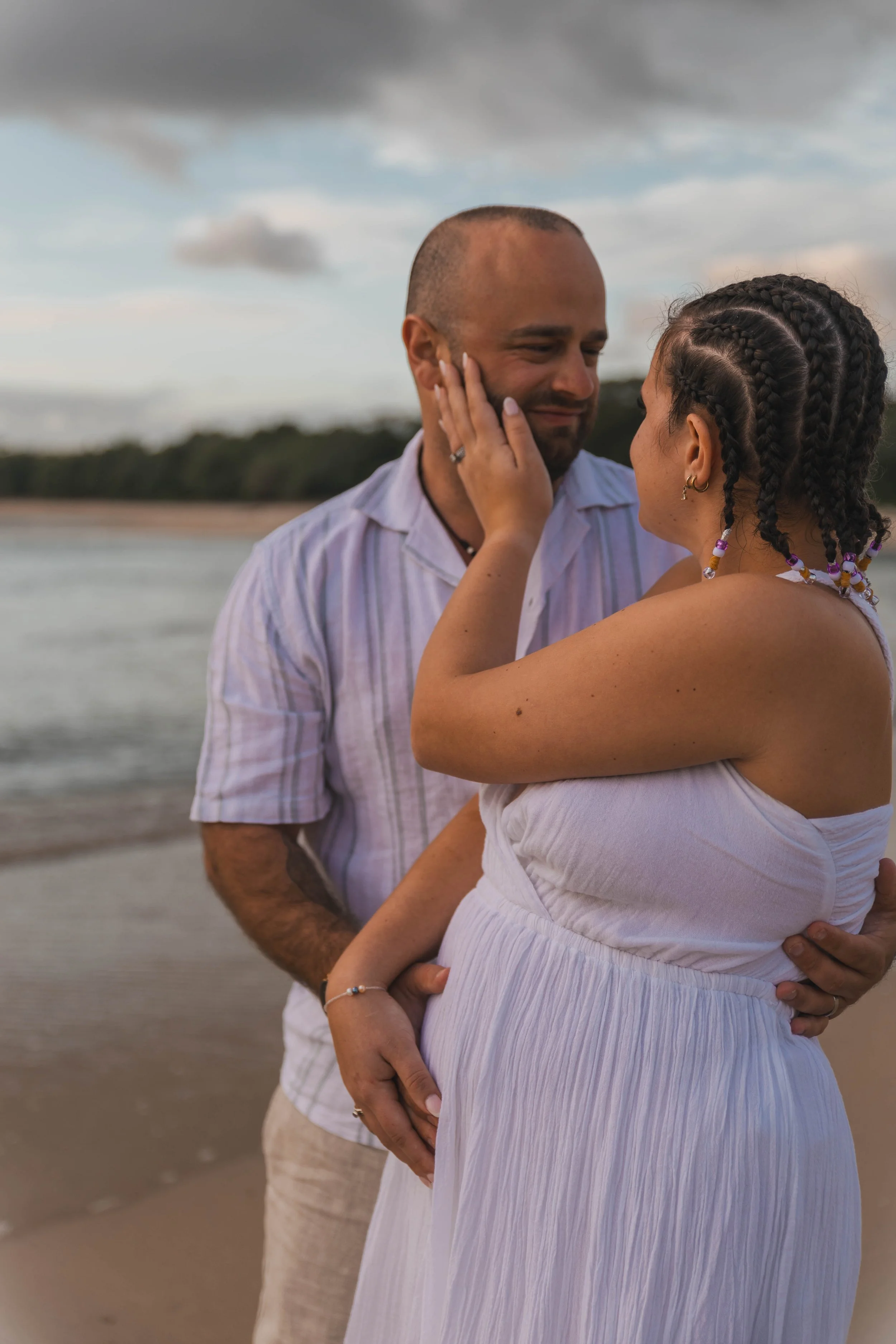 A couple standing on the beach, embracing with ocean in background, cloudy sky overhead.