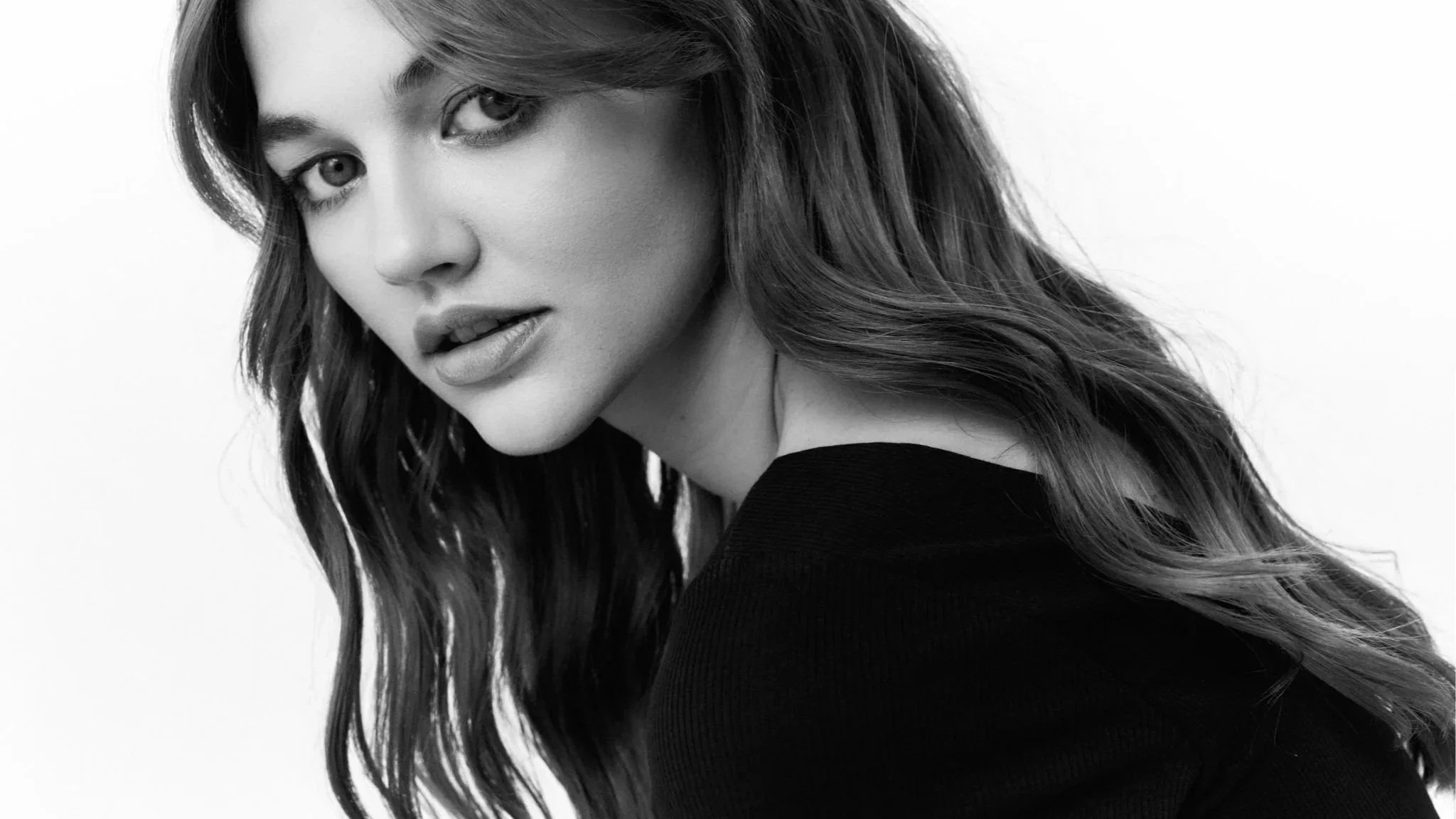 Schwarz-weiße Porträtaufnahme einer jungen Frau mit langem, gewelltem Haar, die eine schwarze Kleidung trägt.