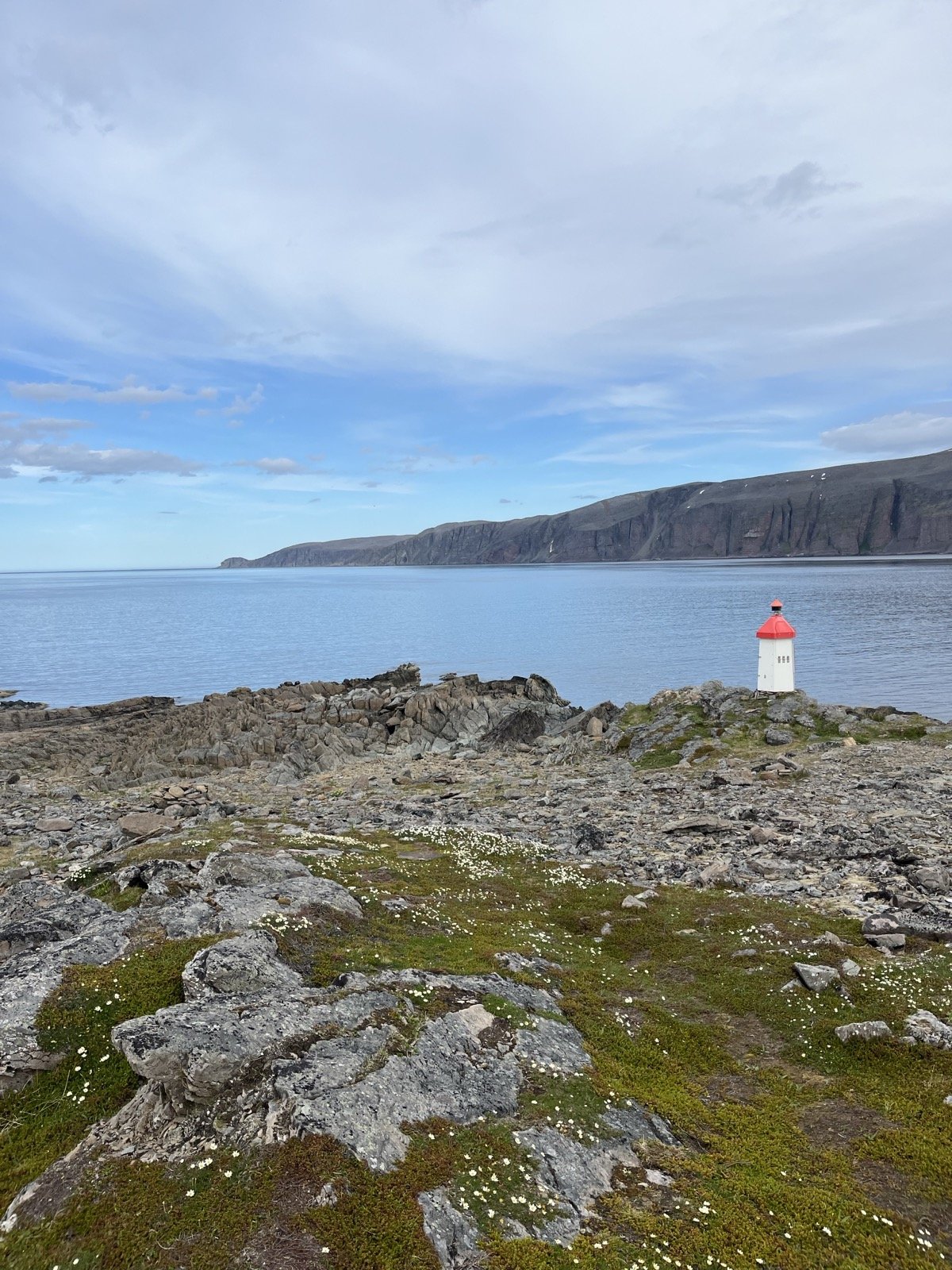 A rocky coastal landscape with a lighthouse with a red roof and white walls, overlooking the Arctic Ocean. Fell cliffs are visible in the background under a cloudy sky.