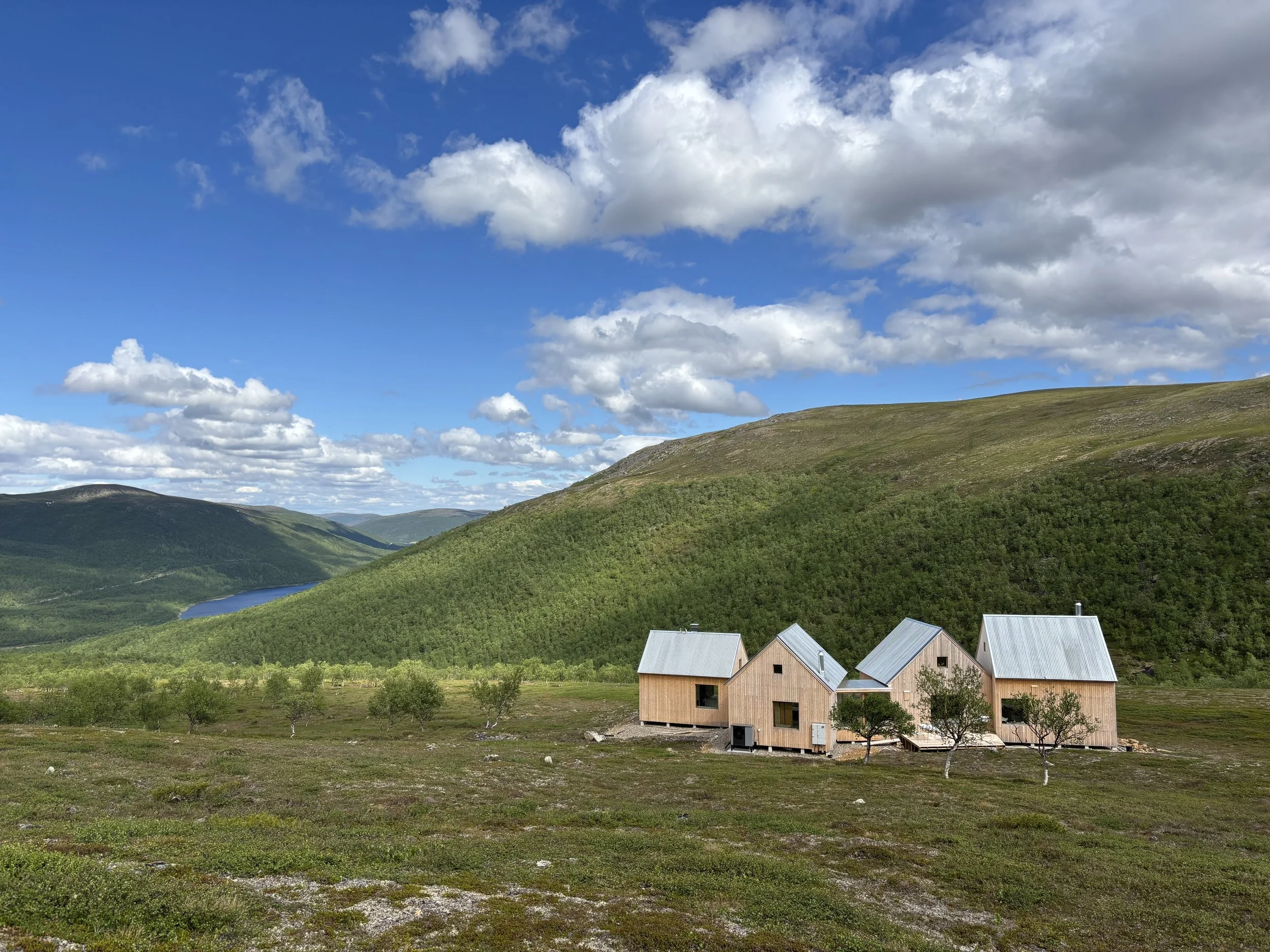 Scenic view of green fells with a glimpse of a river, a few modern wood-clad houses with metal roofs, and a partly cloudy blue sky.