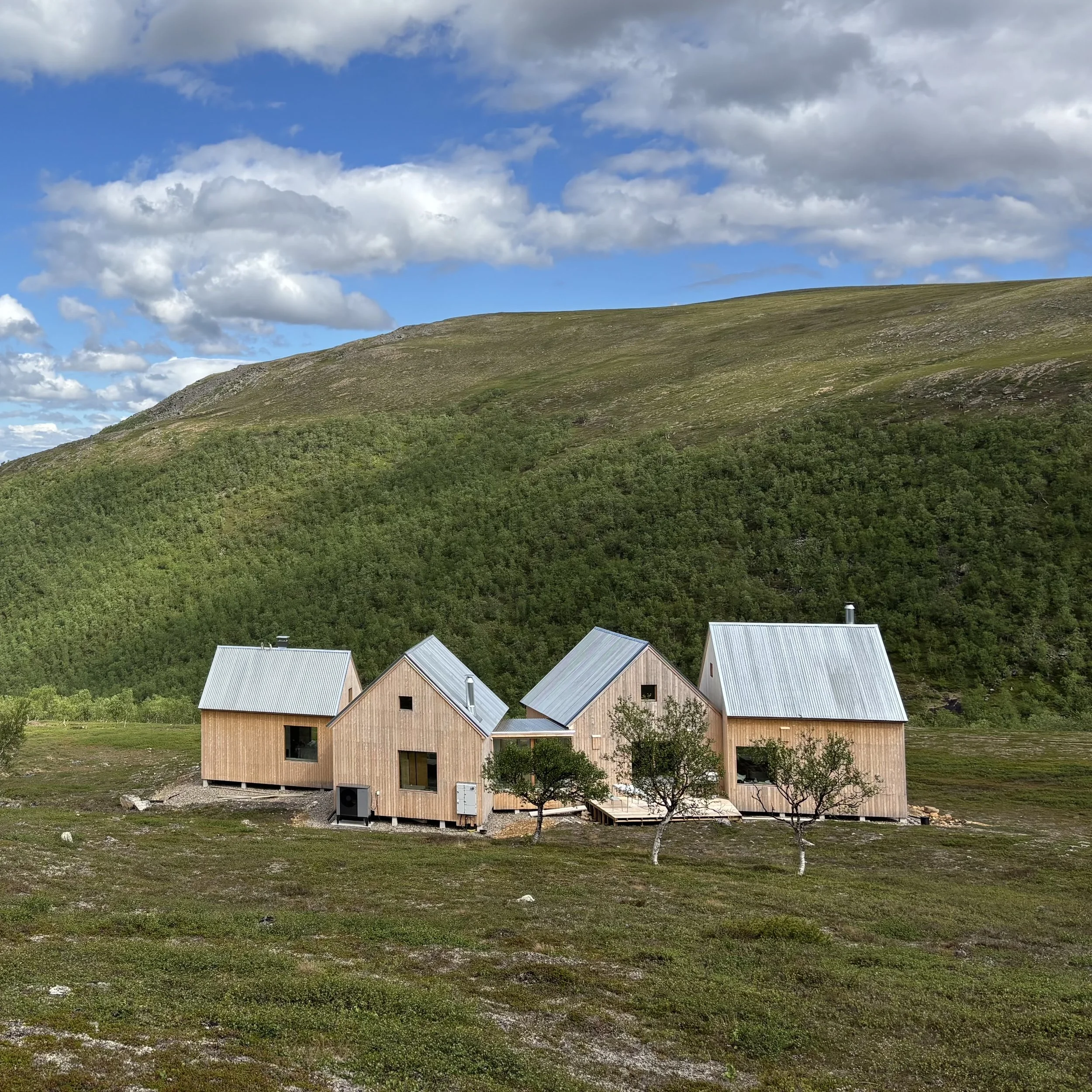 A fell covered with green vegetation and a few Nordic wood-clad cabins with metal roofs at the base of the fell, under a partly cloudy sky.