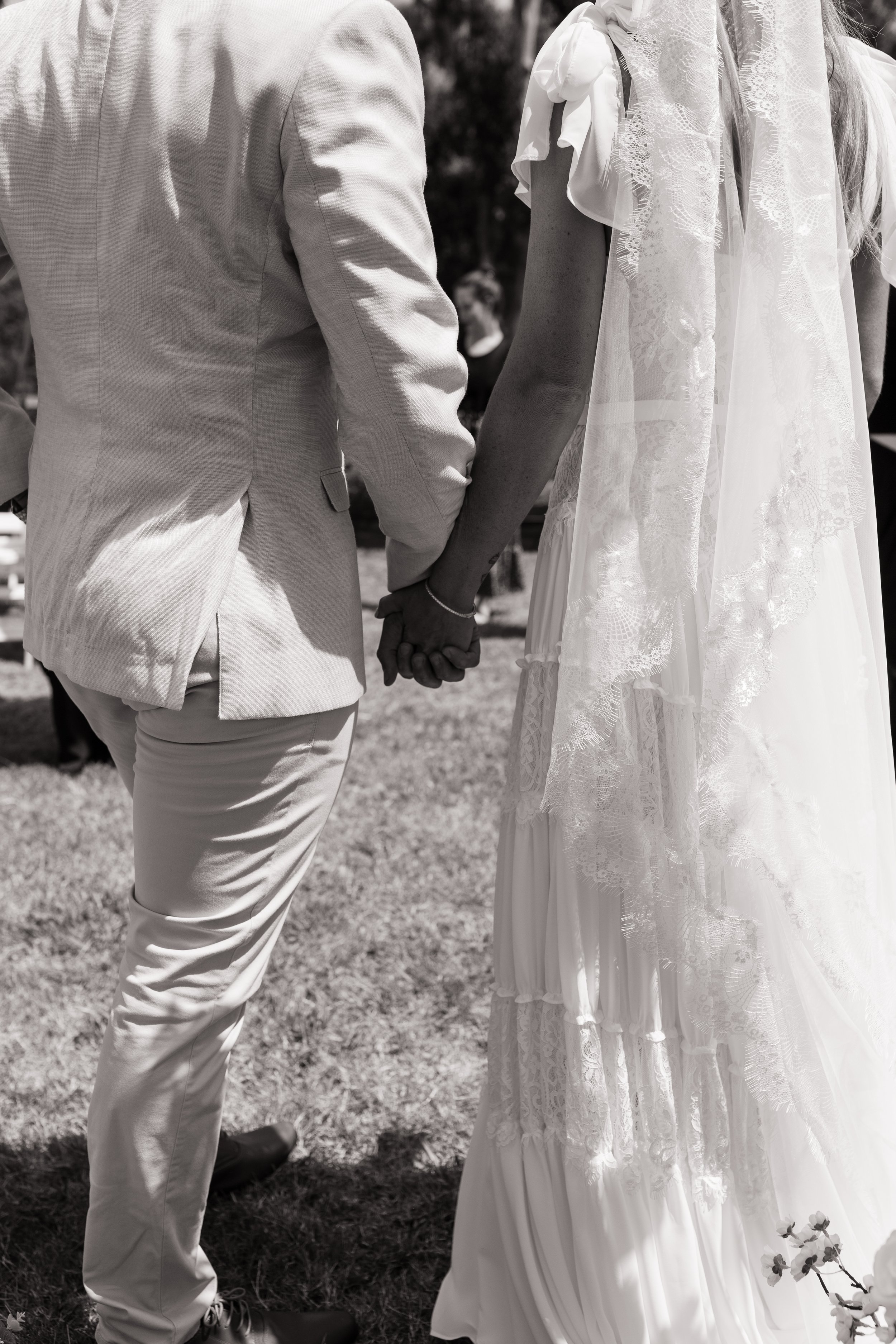 A couple holding hands during a wedding ceremony, with the man wearing a suit and the woman wearing a white lace wedding dress.