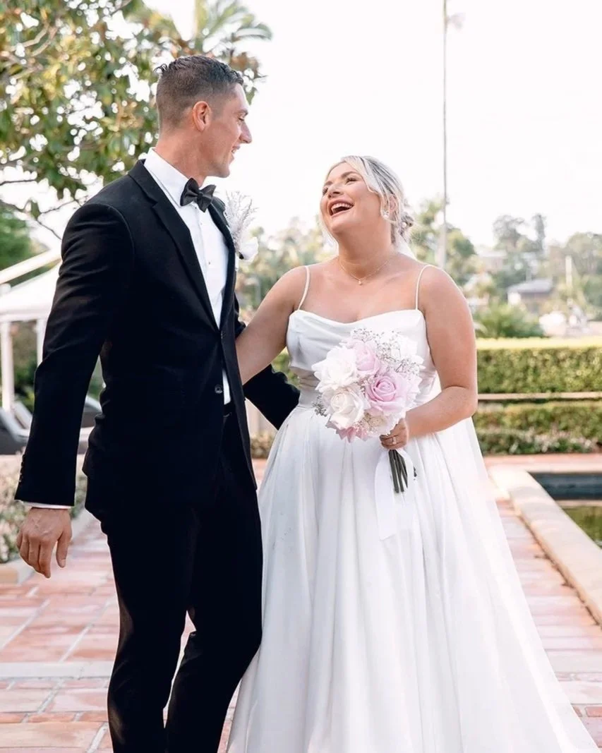 A bride and groom smiling and standing together outdoors during a wedding, with greenery in the background.