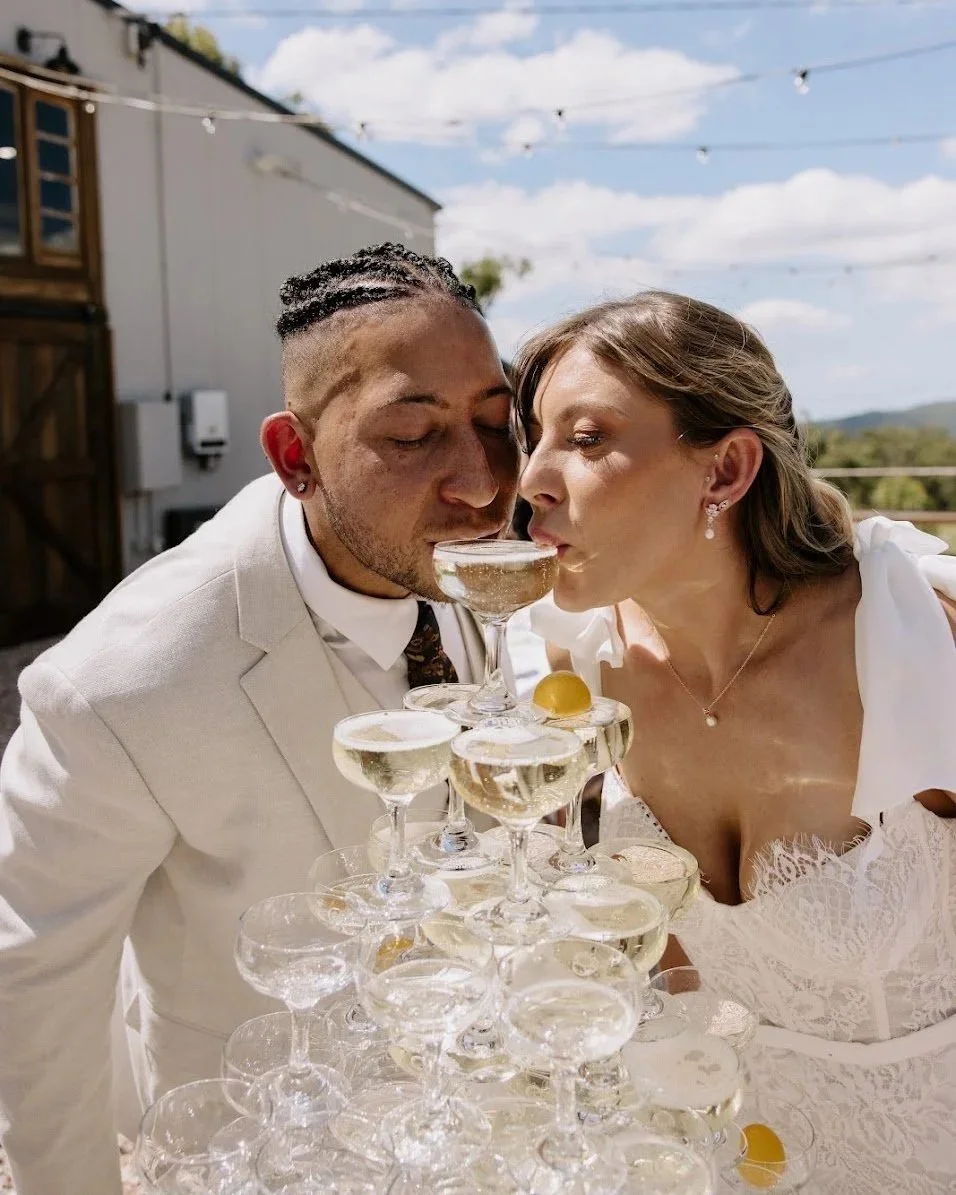 A bride and groom celebrating outdoors sipping from the top of a champagne tower.
