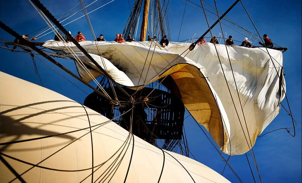 Skeppets segel och rigg set mot en blå himmel, med personer som arbetar under segeln.
