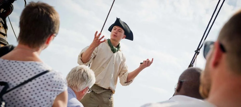 En ung man klädd i historisk uniform, med svart hatt, talar till en grupp personer på en båttur.