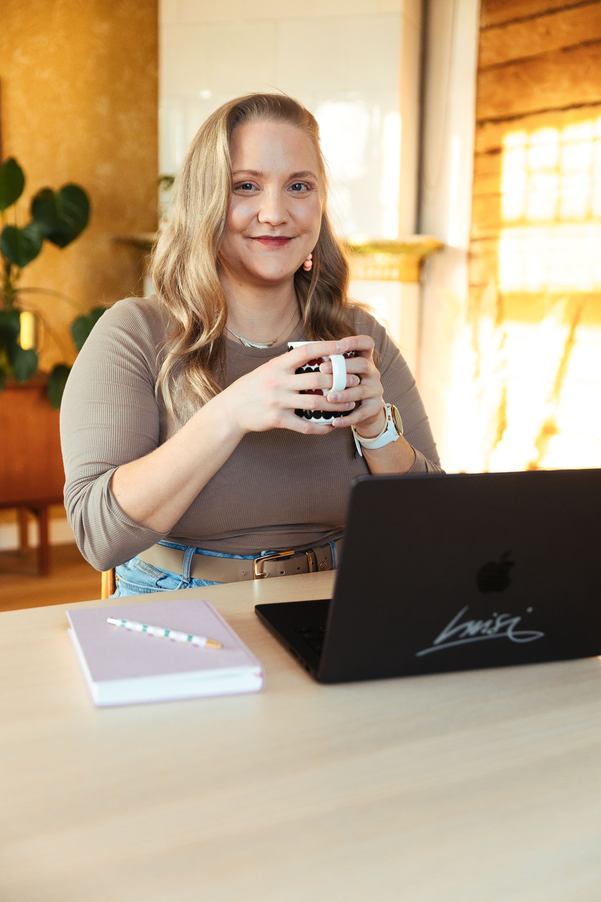 Nuori nainen istuu pöydän ääressä, juo kahvia tai teetä, ja uskoo, että hän on tyytyväinen. Taustalla näkyy ikkunasta valoa ja sisätiloihin kuuluvat kasvit.