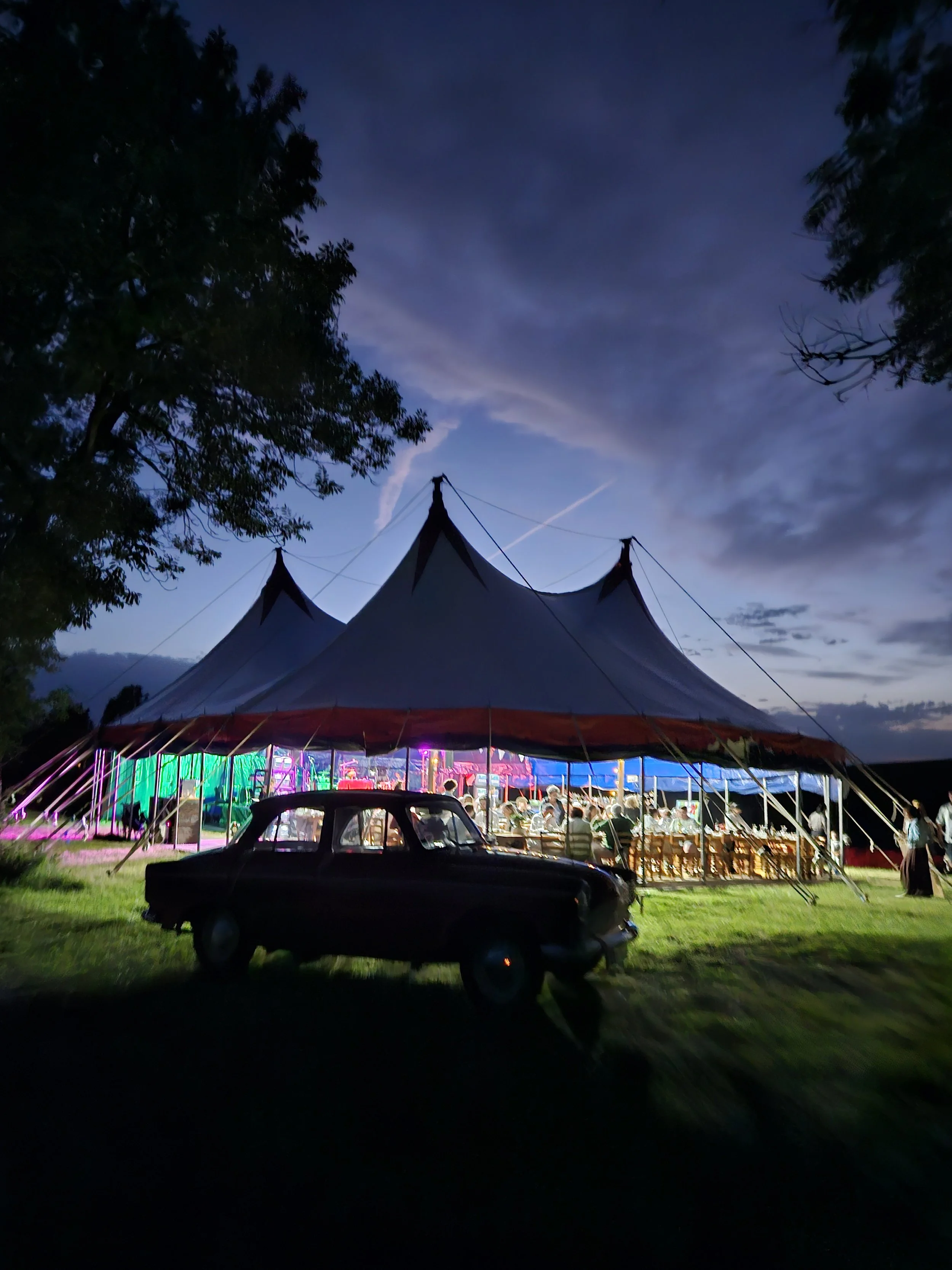 Une tente de cirque éclairée lors d'une soirée, avec des personnes assises à l'intérieur, un arbre à gauche et une voiture ancienne au premier plan, sous un ciel crépusculaire avec des nuages.