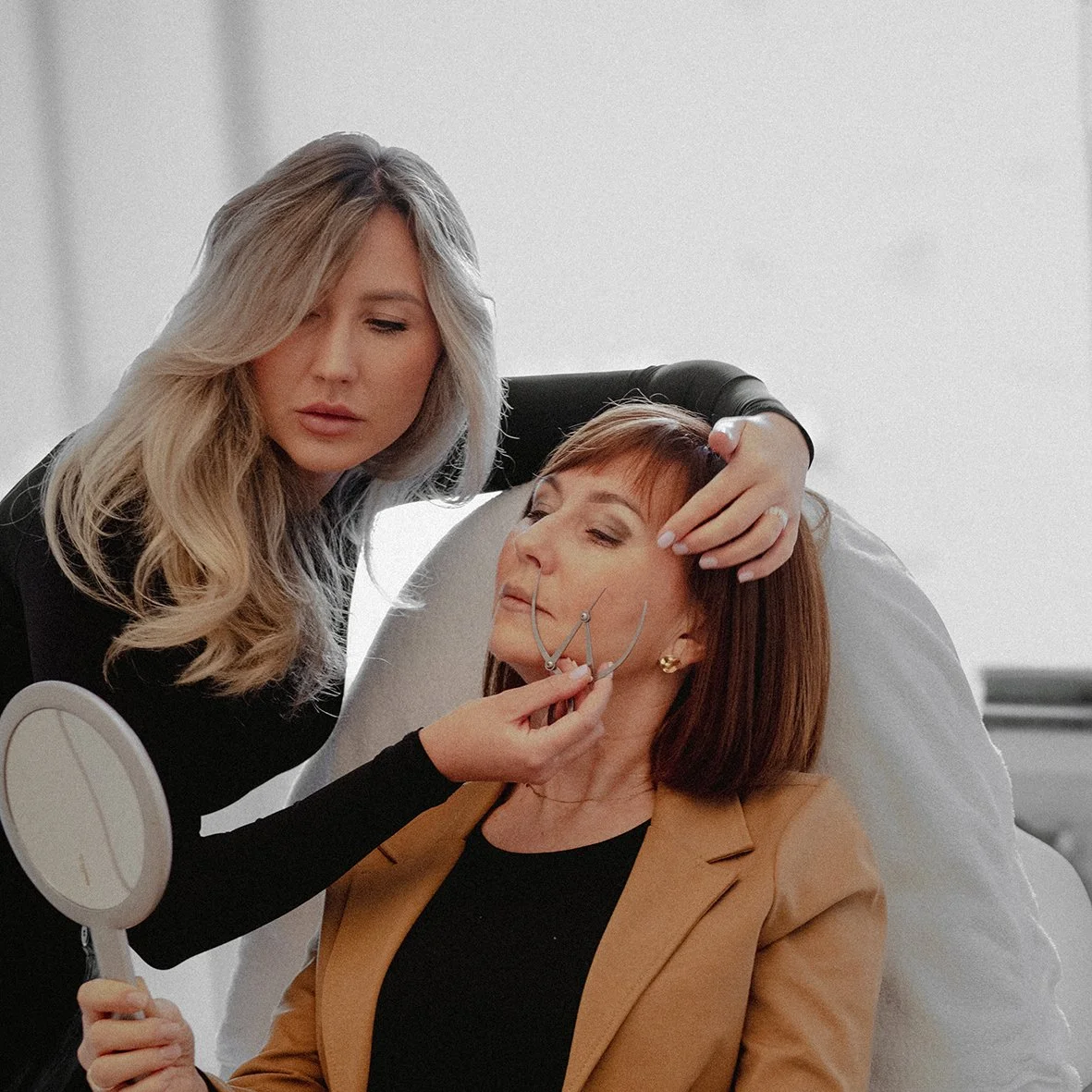 Eine junge Frau überprüft ob es notwendig ist eine Gesichtsbehandlung durchzuführen. Die Kundin hält einen Spiegel und schaut zu.