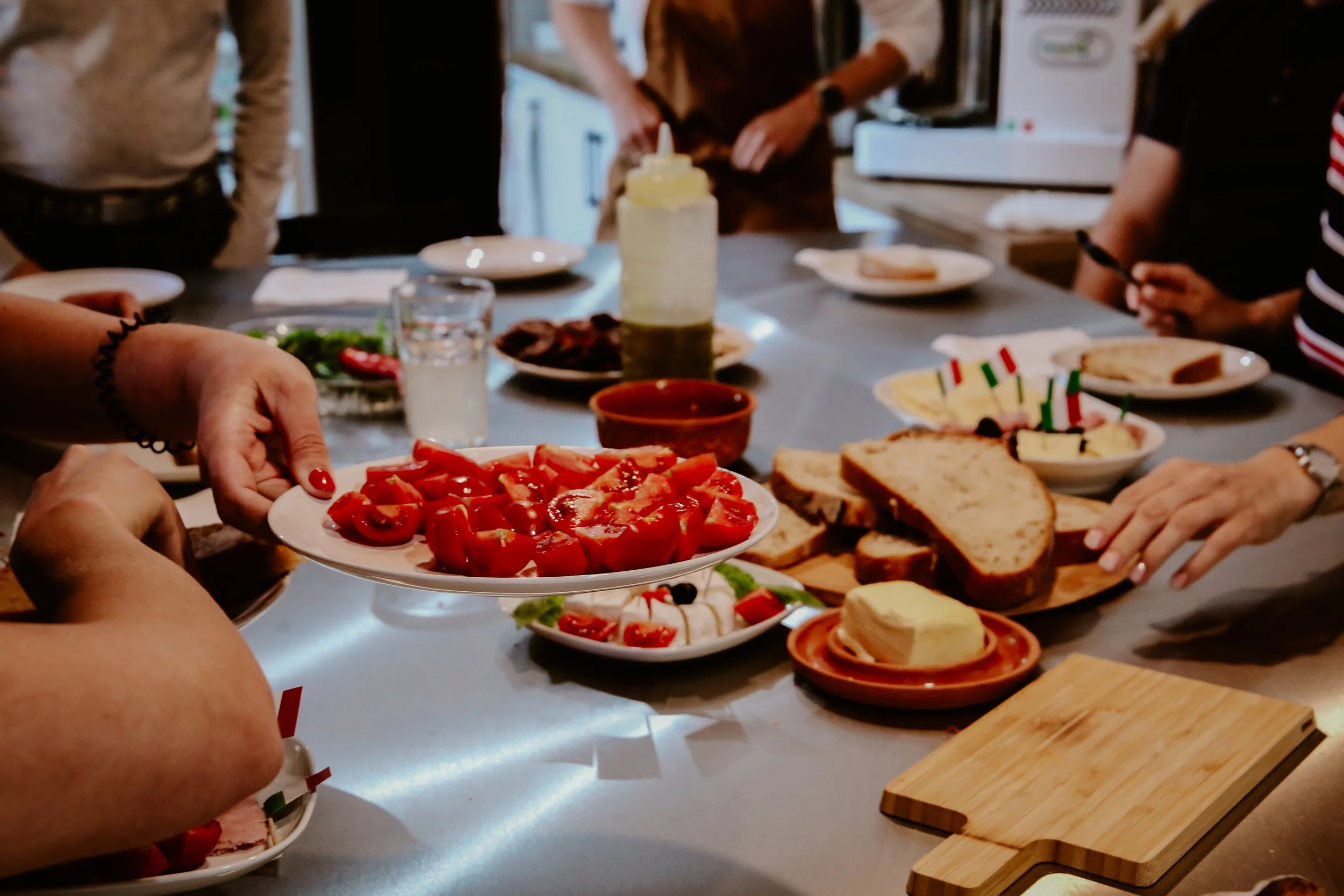 People gathered around a kitchen table with various dishes, including sliced tomatoes, bread, butter, and cheese, preparing for a meal or celebration.