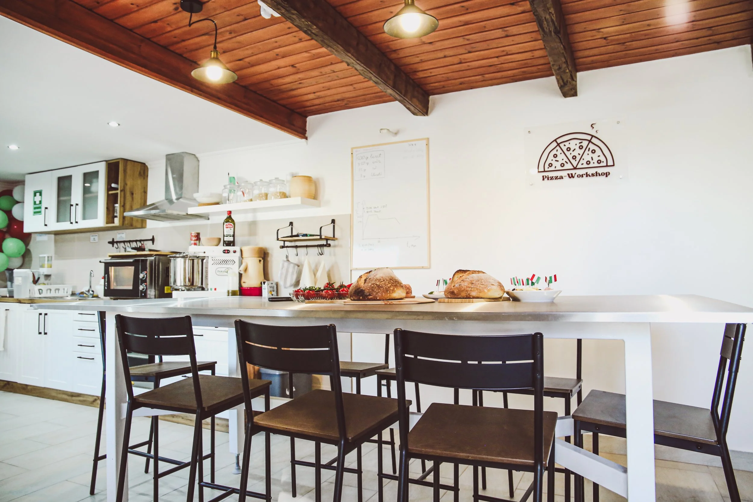 A kitchen with a white island counter, black chairs, and baked goods on the counter. The background has cabinets, a microwave, and kitchen utensils, with a pizza workshop sign on the wall.