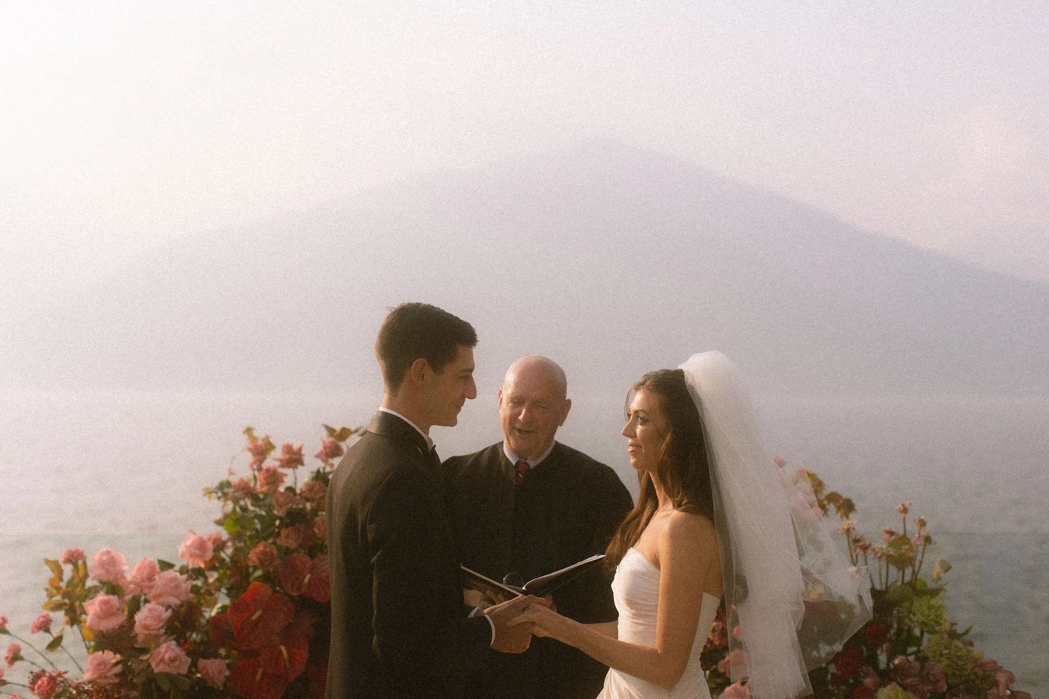 A bride and groom exchange vows during their wedding ceremony outdoors at sunset, with an officiant and pink flowers in the background.