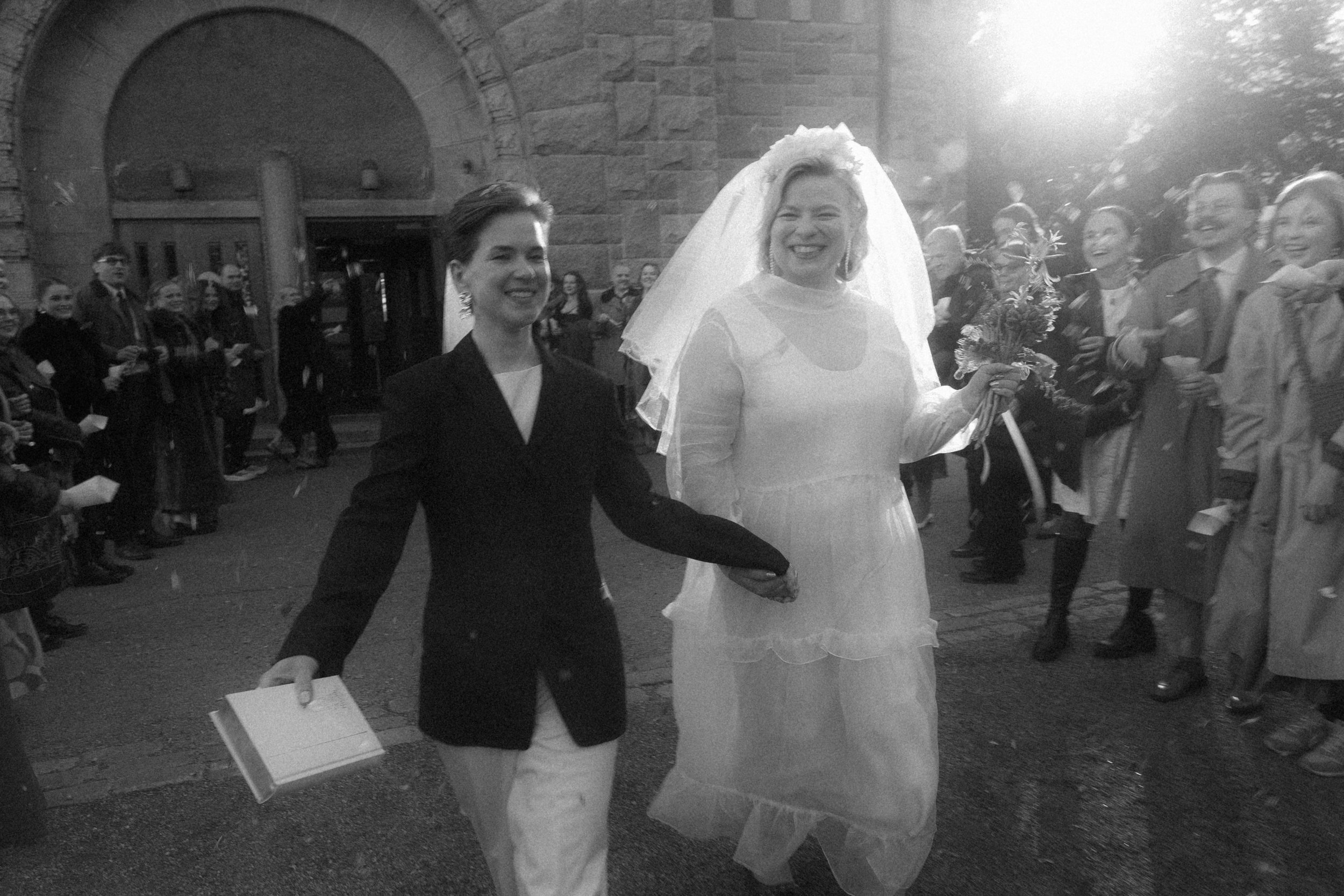 A wedding celebration outside a church featuring two women, one in a formal black jacket and white blouse, and the other in a white wedding dress with a veil, holding a bouquet. They are surrounded by smiling guests under natural sunlight.