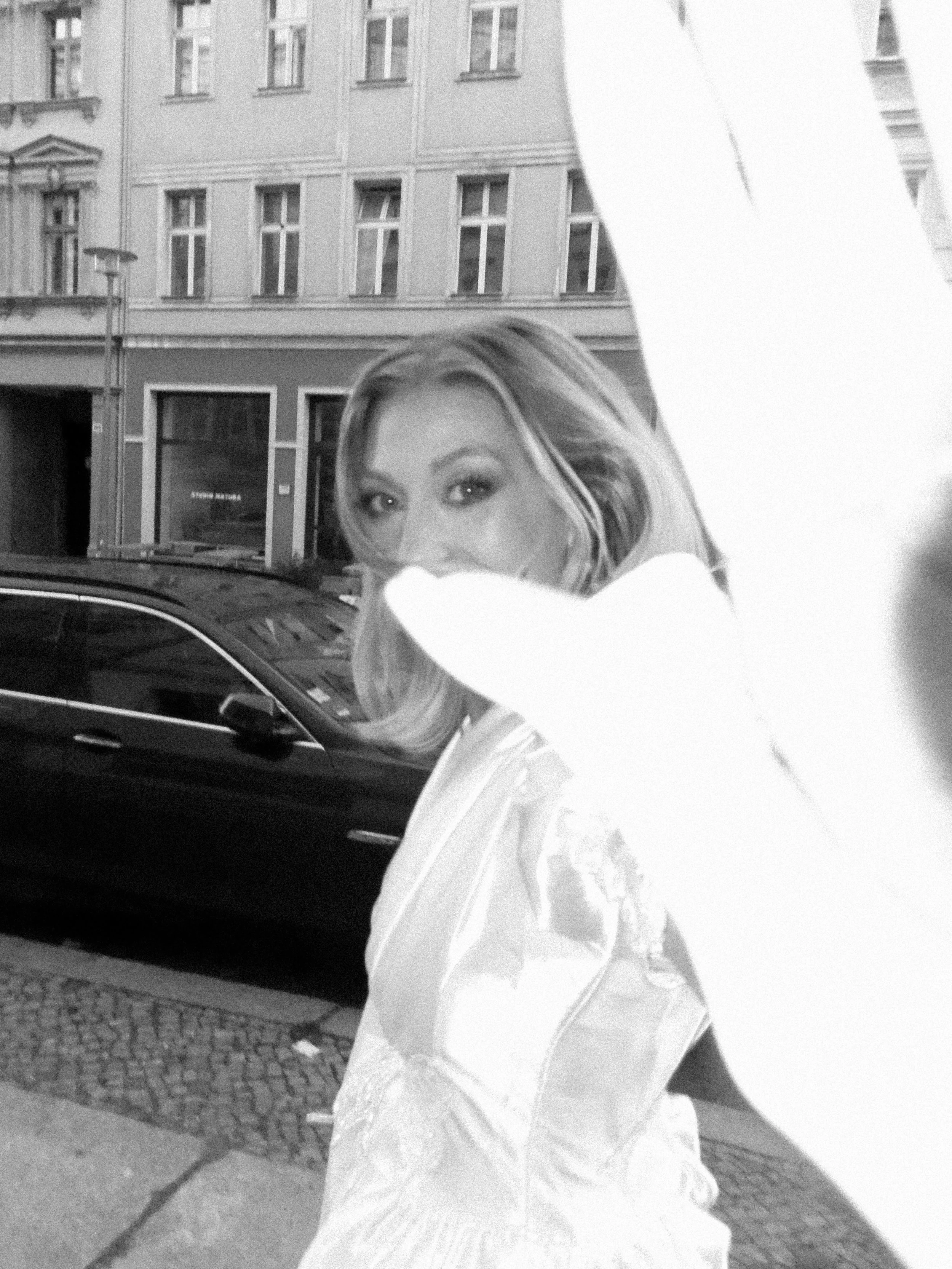 A woman with blonde hair and a white dress, partially obscured by a white coat or blanket, standing on a cobblestone street in front of a black car and a building with multiple windows.