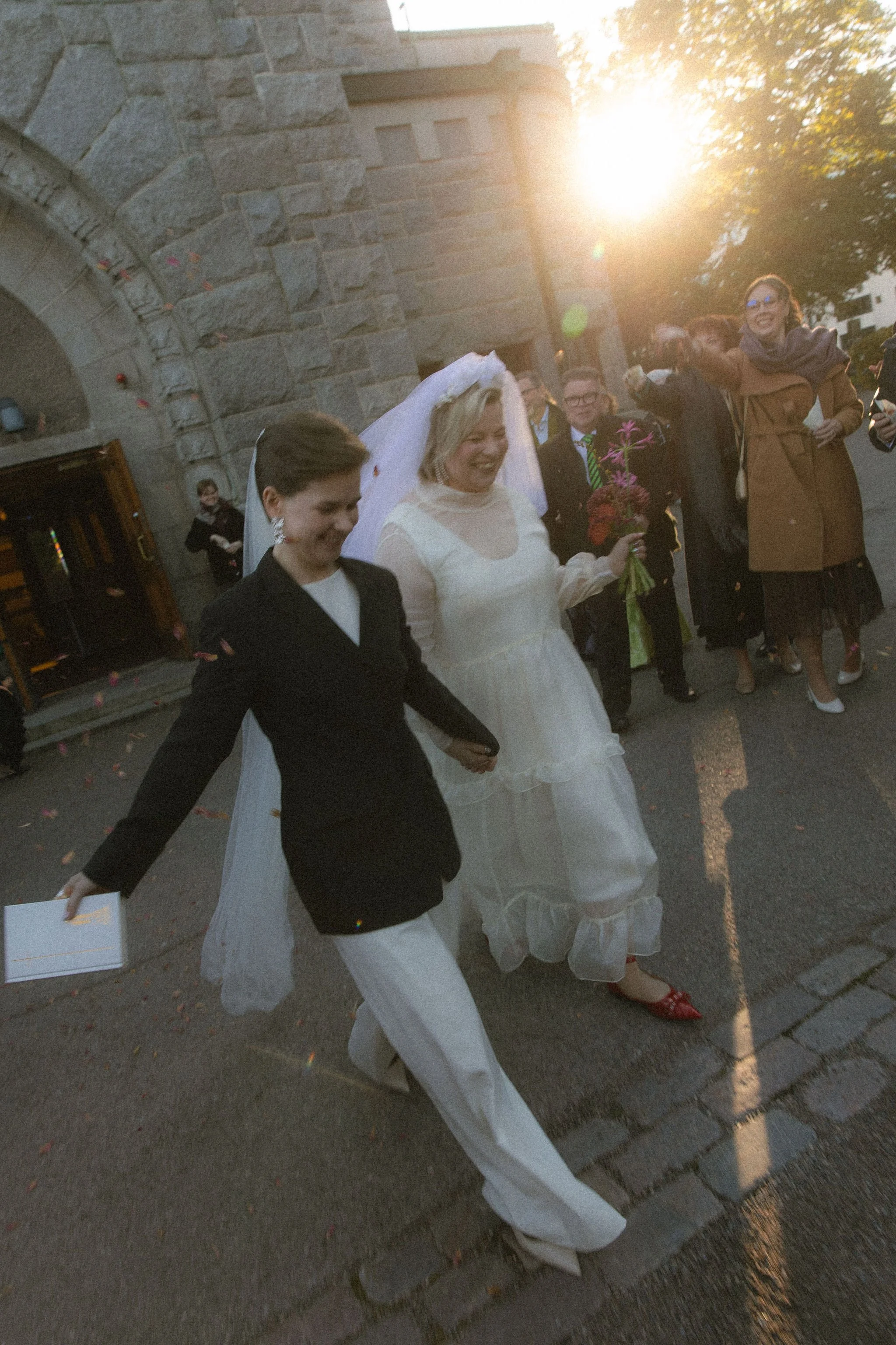 A group of women, including one dressed in a white wedding gown with a veil, walking outdoors on a sunny day near a stone building. They are smiling and appear to be celebrating, possibly after a wedding ceremony.