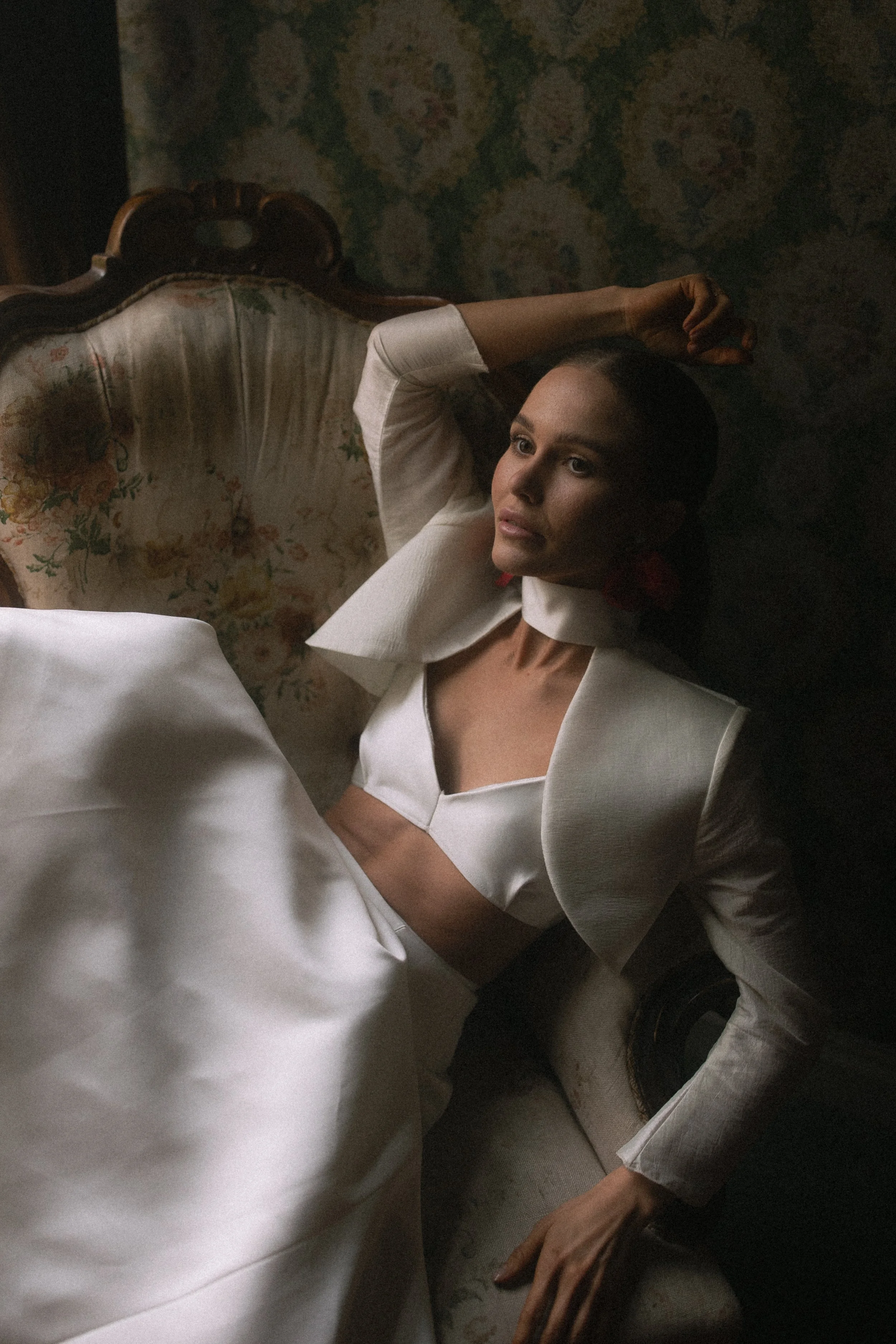 A woman lying on a vintage sofa with floral upholstery, wearing a white blazer and red earrings, with her hand resting on her head and looking thoughtfully to the side in a dimly lit room with patterned wallpaper.