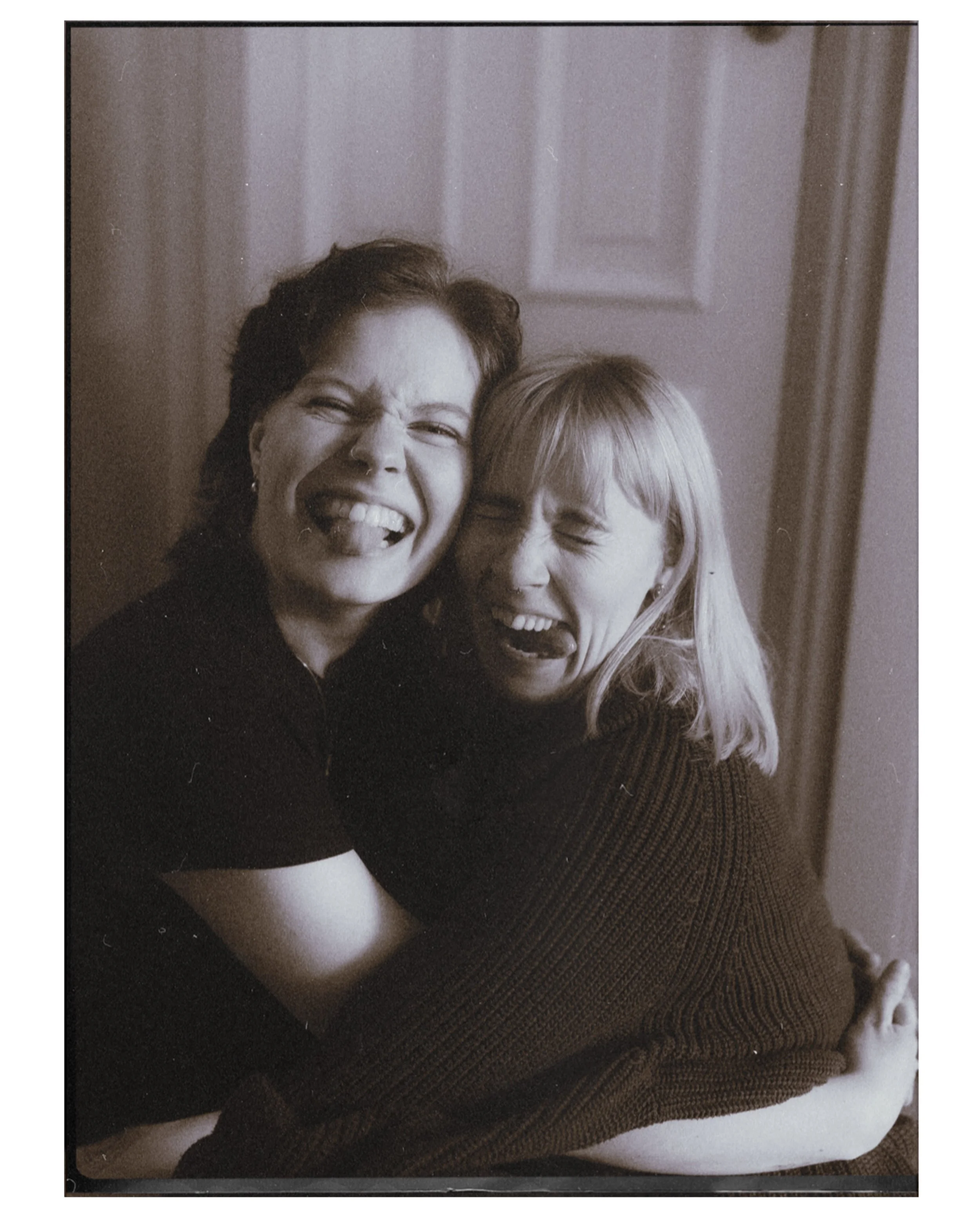 Black and white photo of two women smiling and laughing close together, with one woman wearing glasses and the other with a nose piercing, in an indoor setting.