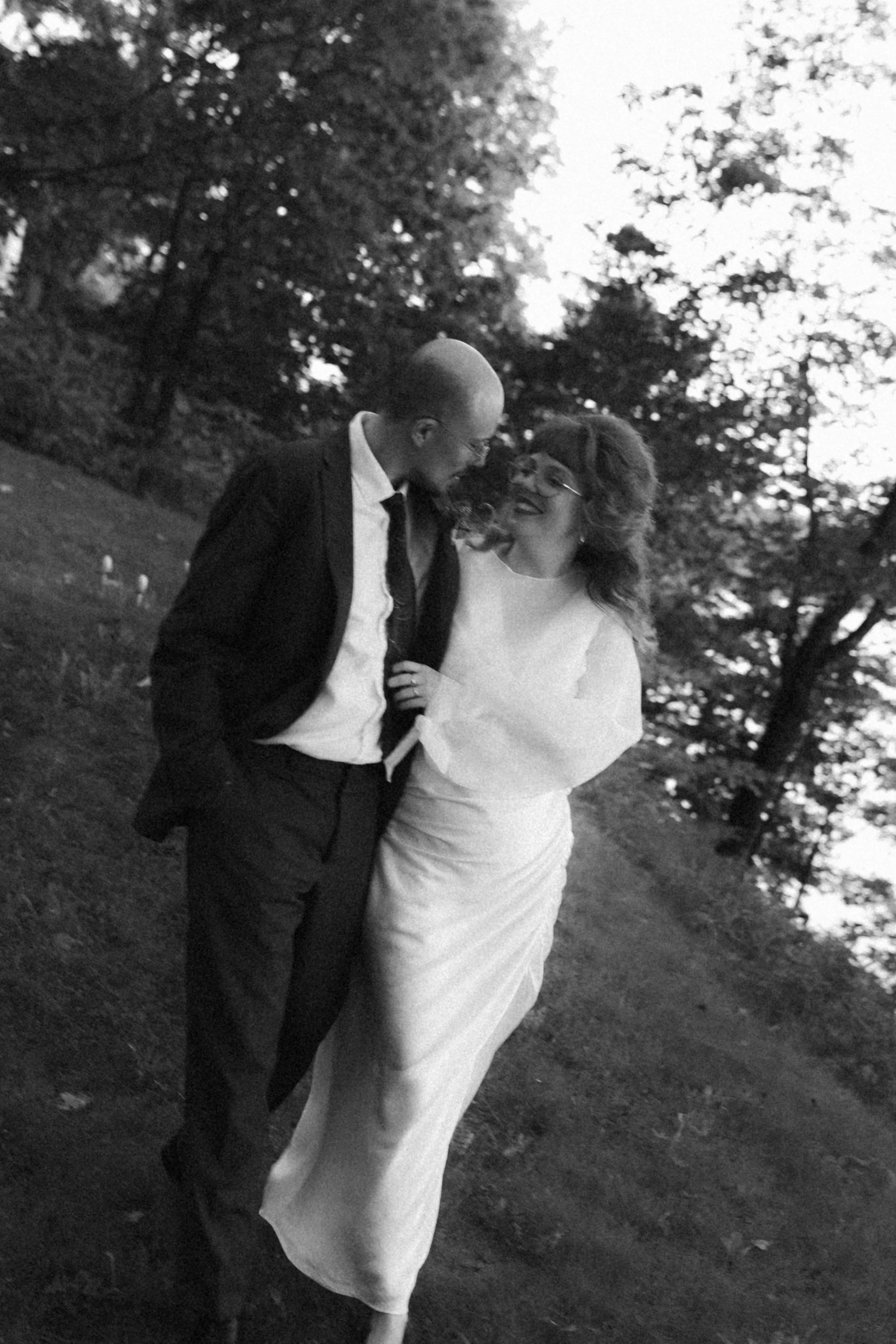 A black and white photo of a man and woman standing close together outdoors surrounded by trees. The man is wearing a dark suit with a white shirt and glasses, while the woman is in a long white dress with curly hair and glasses. They are smiling and
