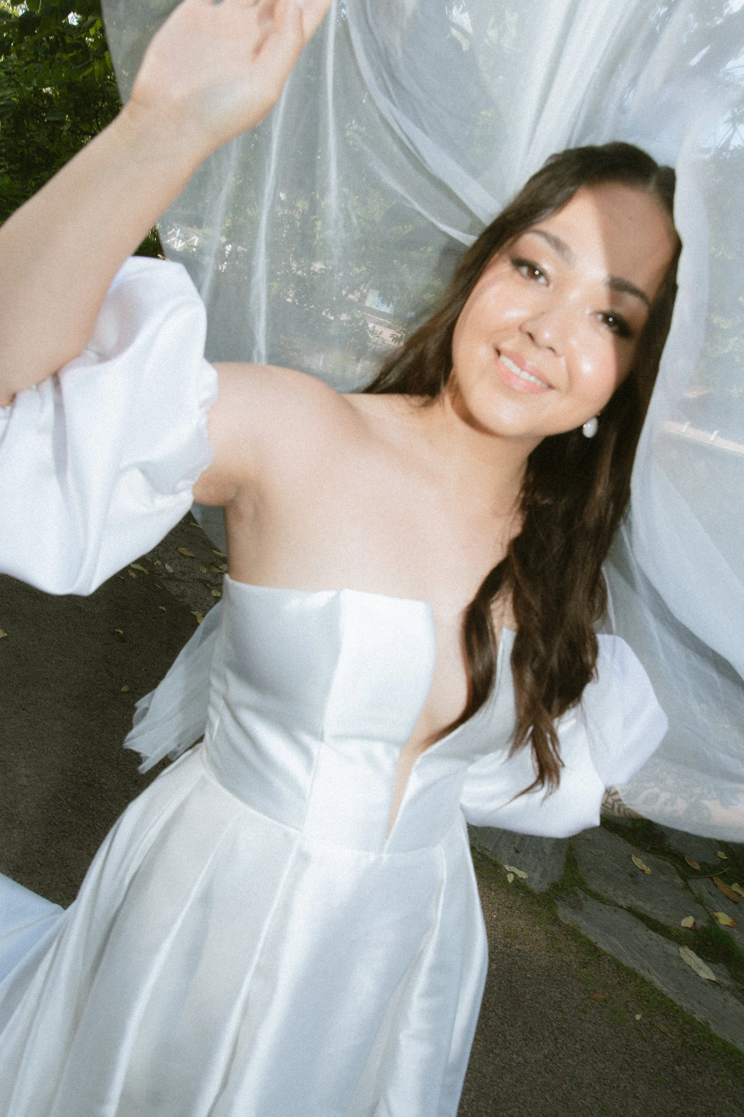A woman with long brown hair in a white strapless dress with puff sleeves, standing outdoors behind a sheer white curtain, smiling at the camera.