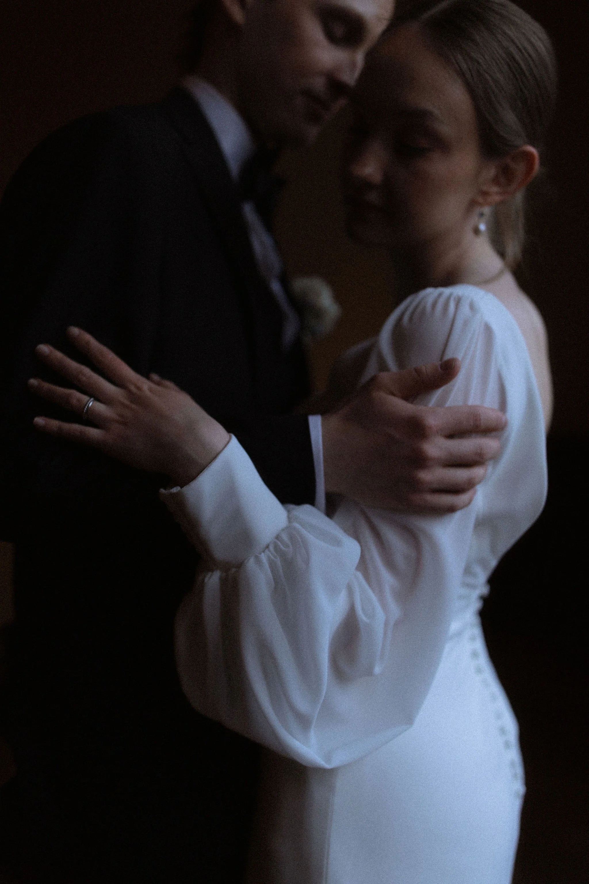 A close-up shot of a couple dancing closely, with their eyes closed and gentle expressions. The man is wearing a dark suit and tie, and the woman is in a white dress with puffed sleeves, both embracing each other.