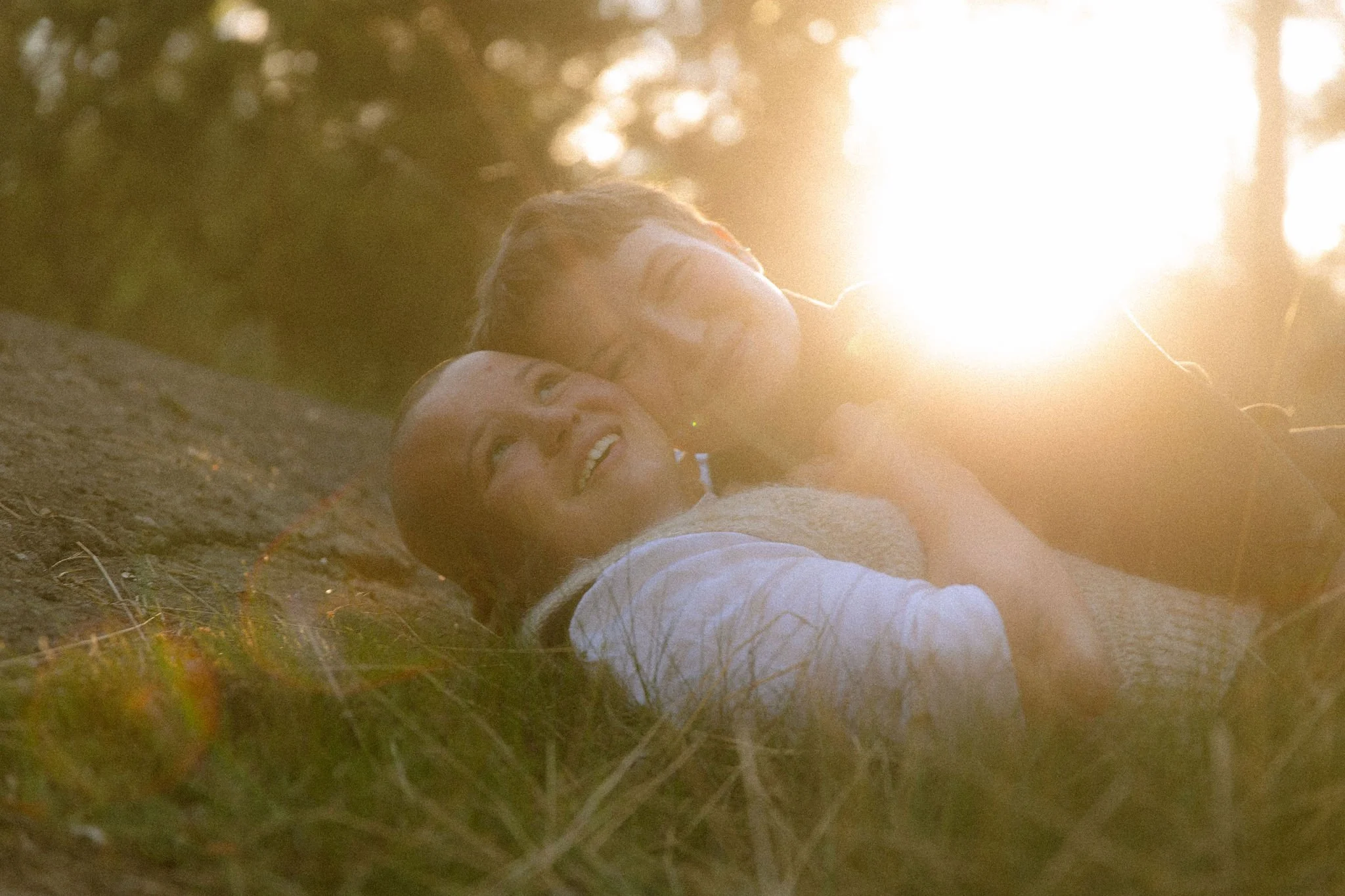 Two people lying on the ground, smiling, with sunlight shining in the background.