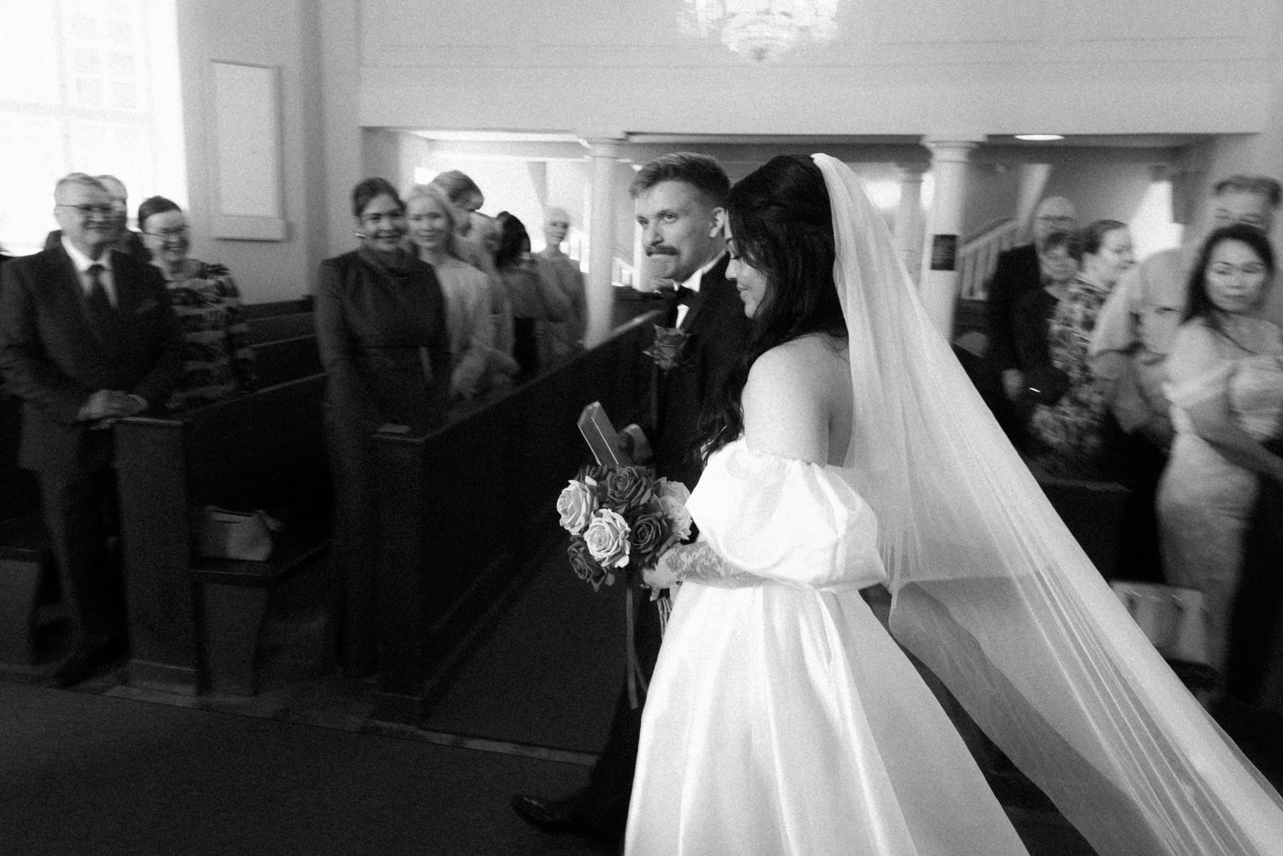 A black and white photo of a bride and groom standing together in a church, surrounded by guests. The bride has long dark hair and is wearing a veil and off-shoulder wedding dress, holding a bouquet of roses. The groom has short hair, a mustache, and is dressed in a tuxedo with a bow tie. The guests are standing, smiling, and looking toward the couple.