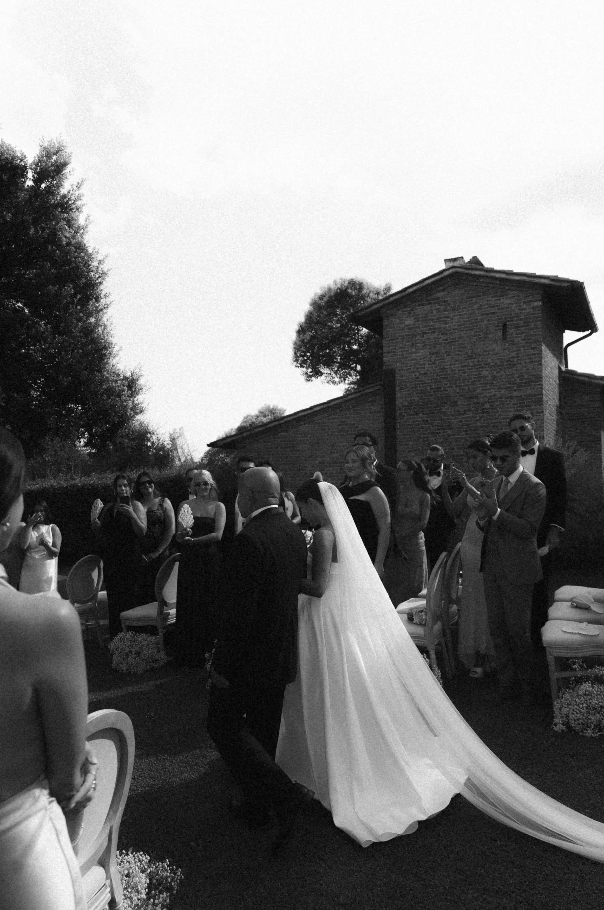 A wedding ceremony outdoors with guests in formal attire, a bride in a white gown, and a groom in a dark suit, surrounded by chairs and some trees and a brick building in the background.