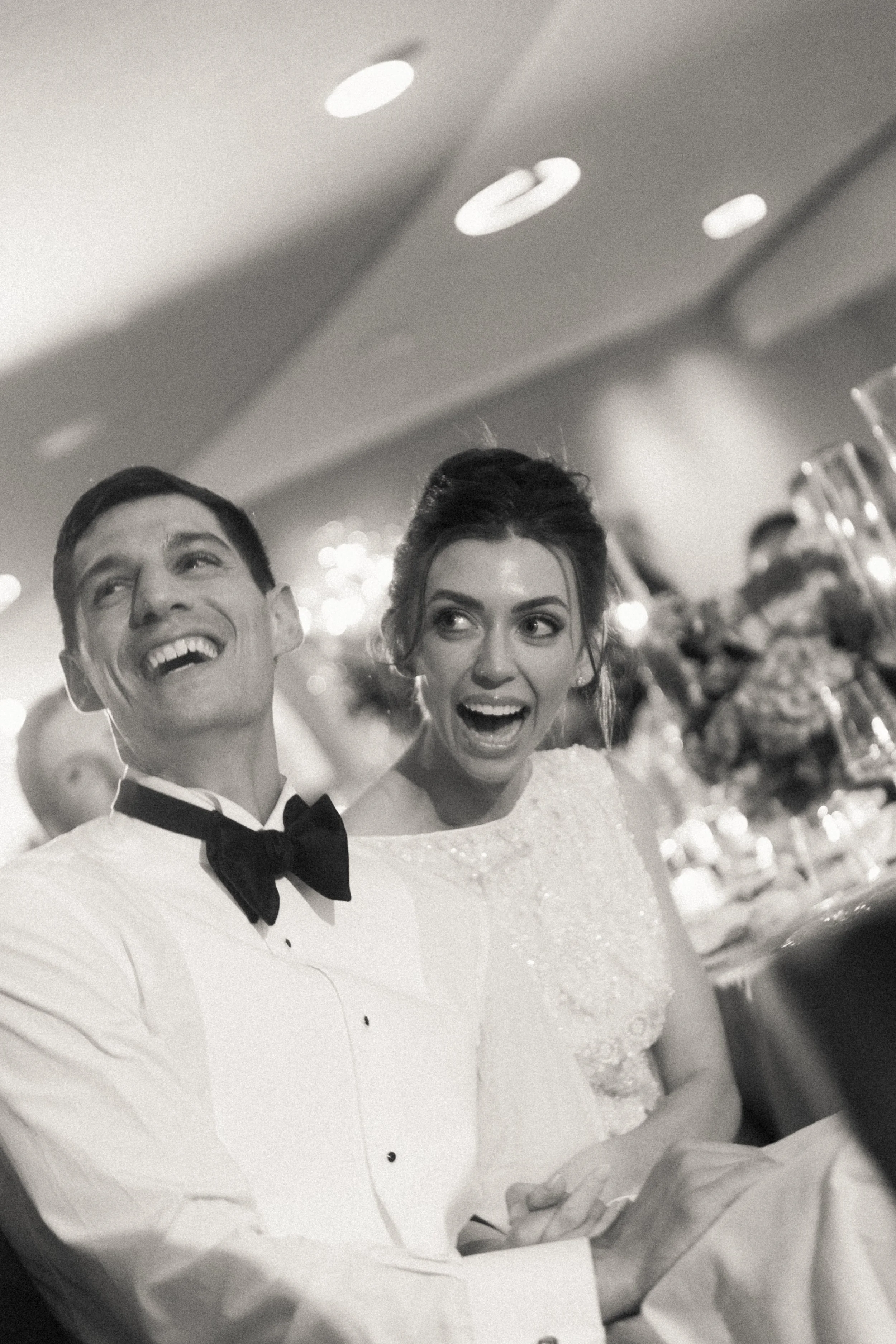 A black and white photo of a smiling man and woman dressed formally, with the man in a tuxedo and the woman in a wedding dress, celebrating at a wedding reception.