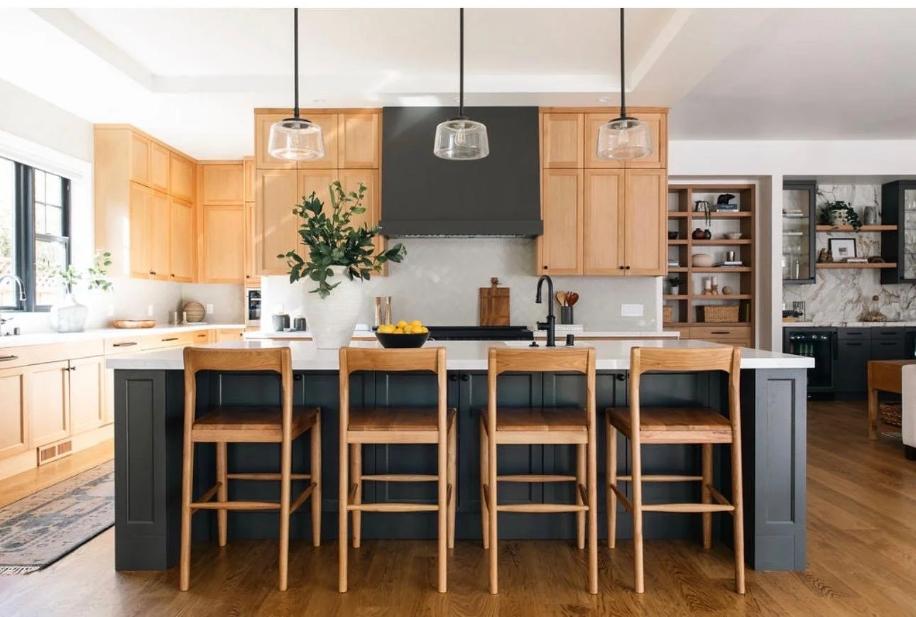 Modern kitchen with wooden cabinets, a navy blue kitchen island, and four wooden barstools. There are pendant lights hanging above the island, a large plant in a white vase, and a bowl of lemons on the counter. The space features open shelving with d
