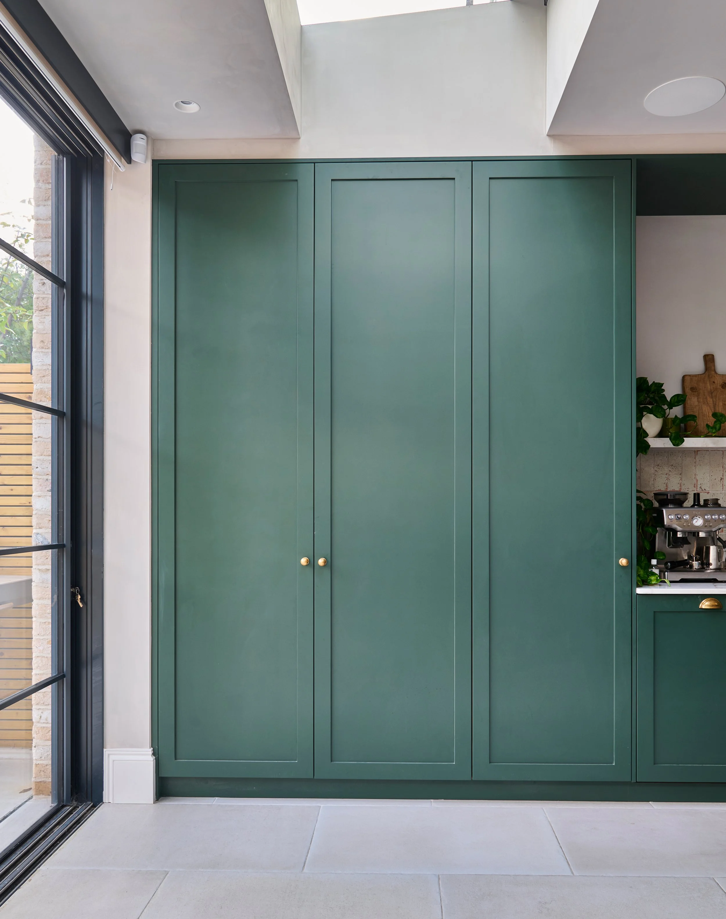 View of a green cabinet with gold knobs in a modern home kitchen, adjacent to a glass door showing an outdoor porch area with wood fencing and plants.