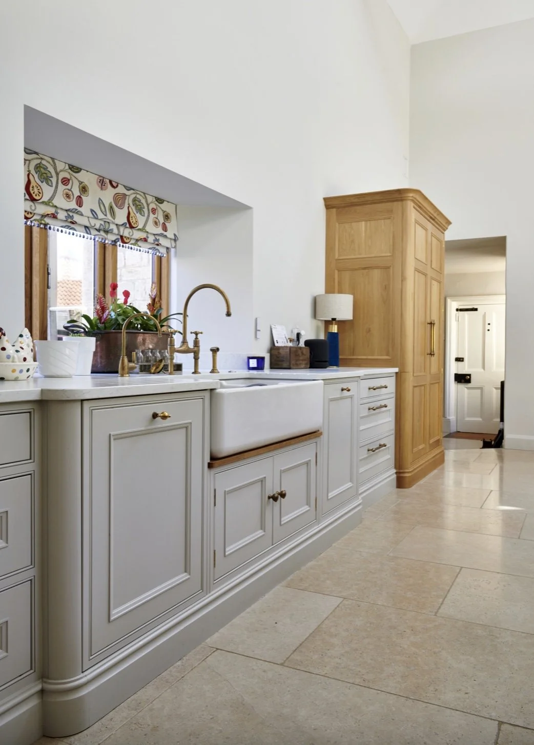 A kitchen with white cabinetry, a large farmhouse sink, brass faucet, window above the sink with a patterned valance, wooden cabinet, and beige tiled floor.