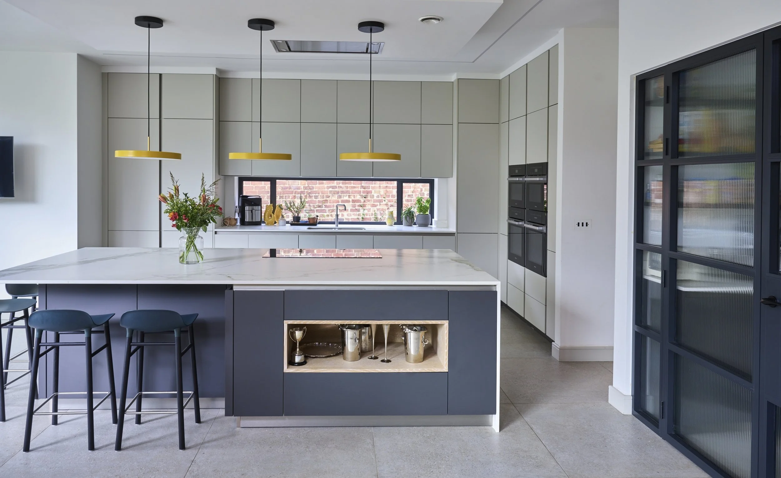 Modern kitchen with light gray cabinets, a large white marble island with a built-in shelf holding trophies and glasses, black bar stools, a window with brick wall outside, yellow pendant lights, and black framed glass door.