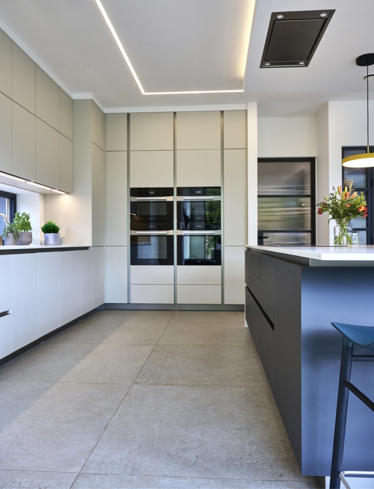 Modern kitchen with white and beige cabinets, a kitchen island with black drawers, built-in ovens, a window with potted plants, and a flower arrangement on the island.
