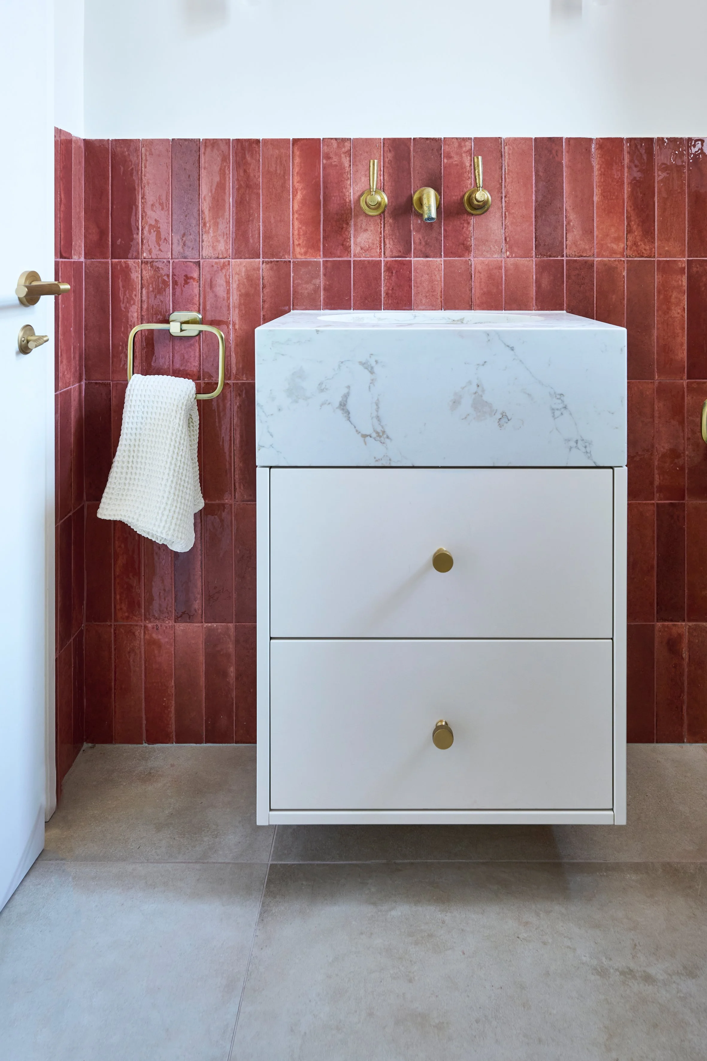 Modern bathroom vanity with a white marble sink, red tiled wall behind, brass fixtures, and beige tiled flooring.