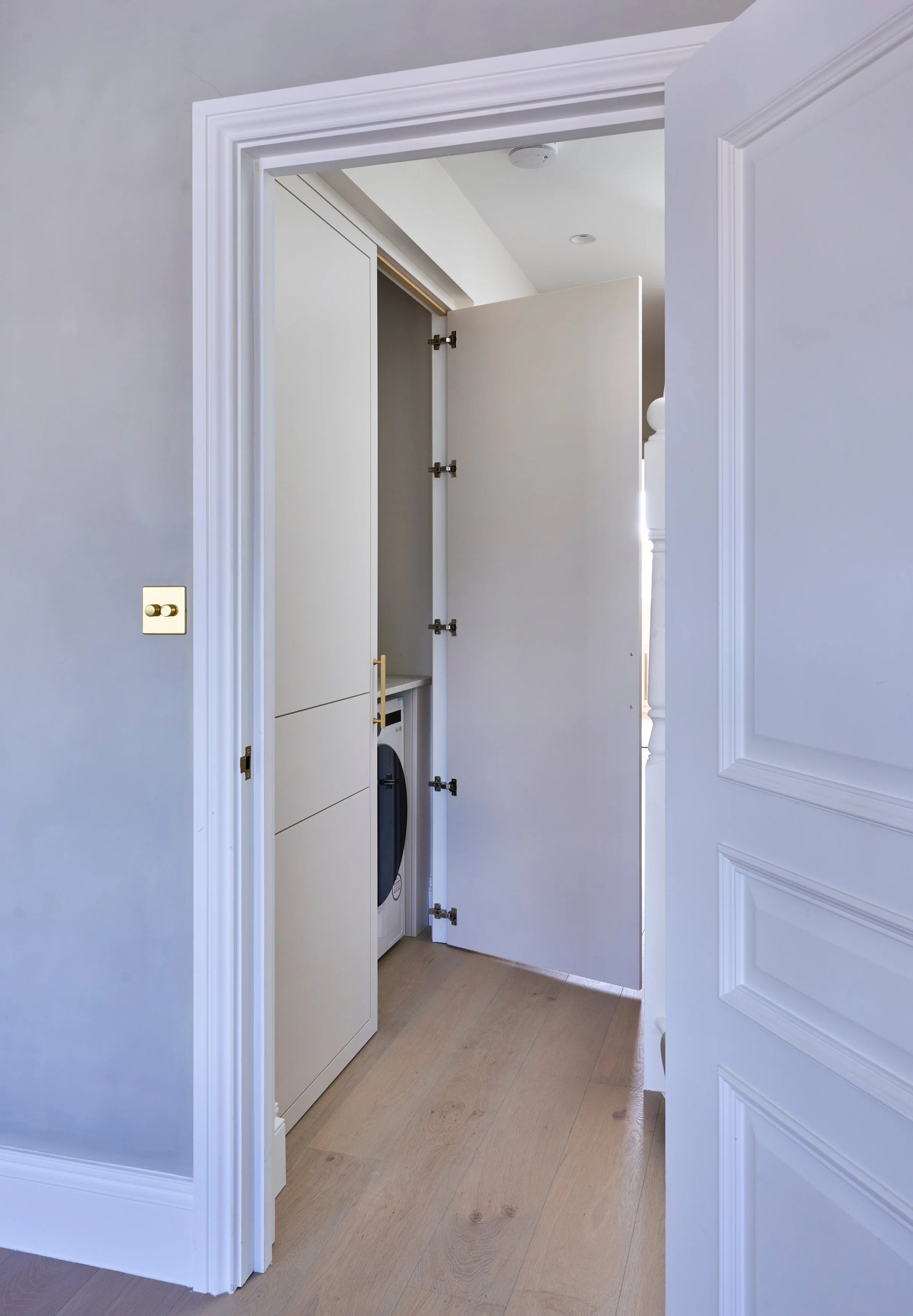 A white door is open, revealing a closet with shelves and a washing machine inside. The room has light-colored walls and flooring, with crown molding and a light switch on the wall.