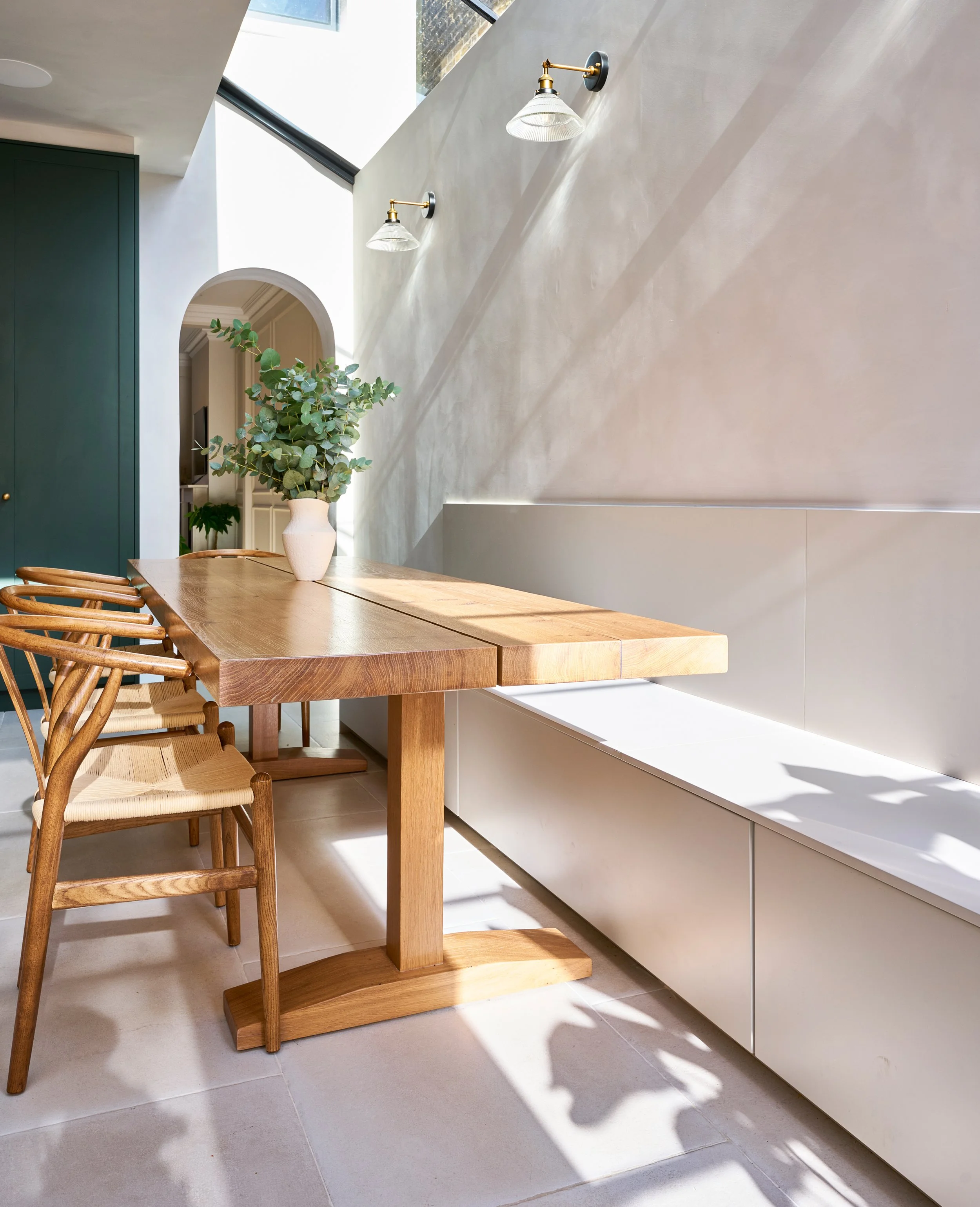 A modern dining area with a wooden table and chairs, a potted plant, white walls, and two wall-mounted light fixtures. Natural sunlight streams through a large skylight.